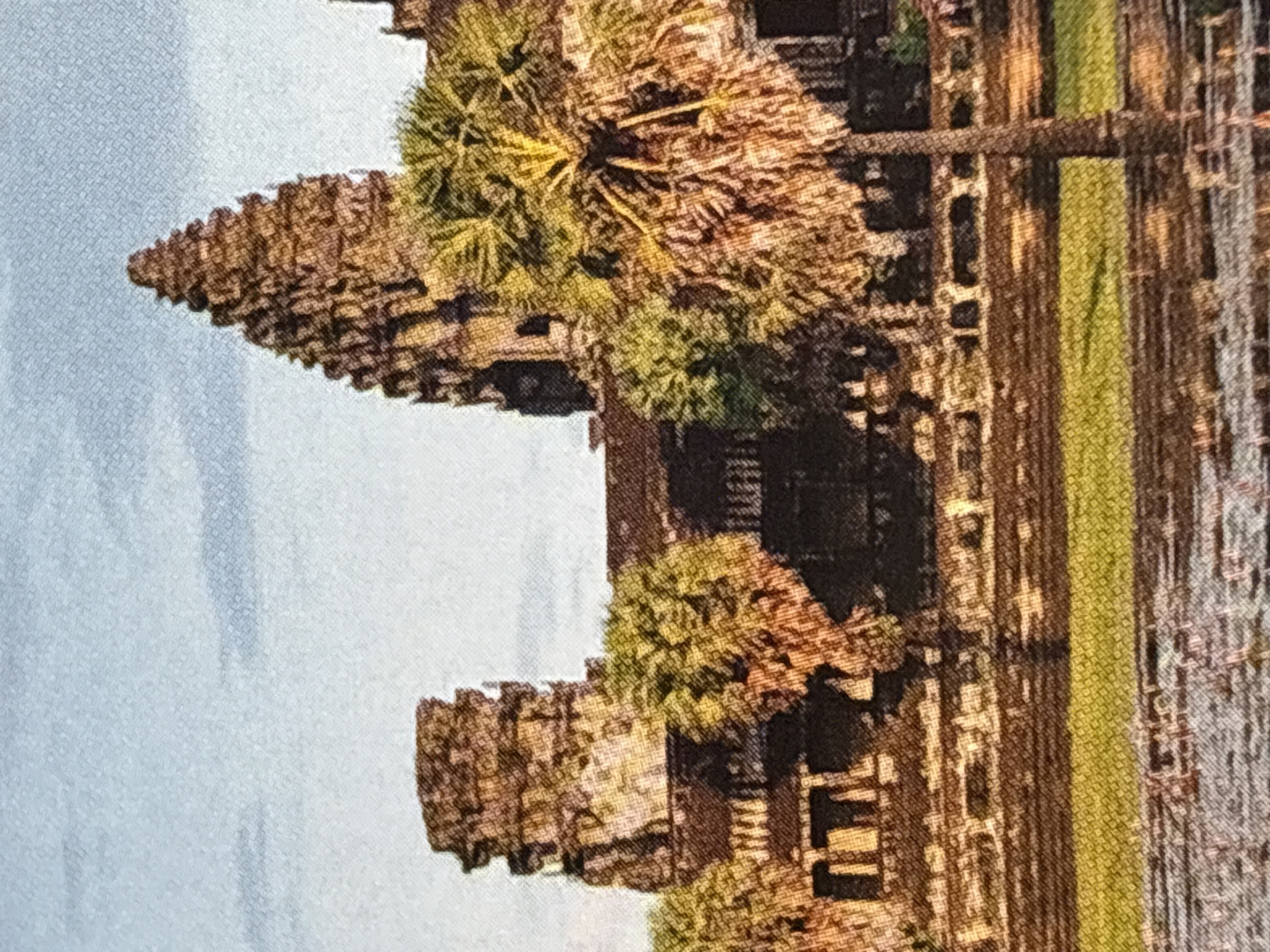 The image depicts a wide shot of Angkor Wat, a temple complex in Cambodia. In the foreground, a body of water is visible with reflections. Behind the water is a grassy area and then the stone structure of Angkor Wat itself. The temple has multiple towers, with the central, tallest tower prominently featured. There are also trees, including palm trees, in front of and around the temple. The sky above is a clear blue with some faint clouds. The image is a photograph, likely taken during the day. There are no people or animals visible in the image. The image has a slightly grainy texture, suggesting it may be a reproduction or a lower-resolution photograph. The location is not in Chelmsford, United Kingdom.