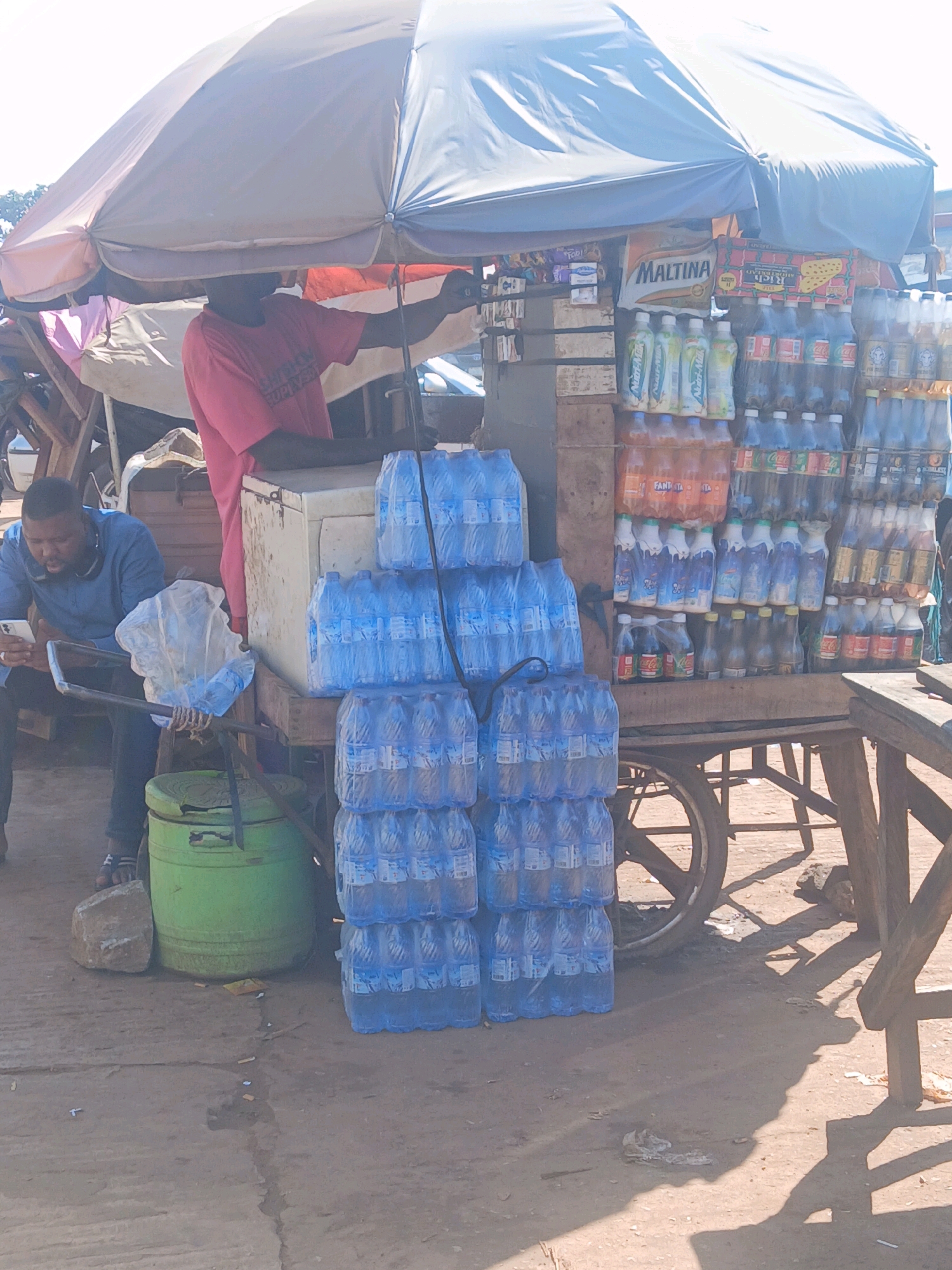 A street vendor in Mokwa, Nigeria operates a cart filled with bottled water and other beverages. The vendor, a man wearing a pink t-shirt, is partially obscured by a large umbrella. In the foreground, another man, dressed in a blue shirt, sits and uses his phone, holding a plastic bag. The cart is laden with stacks of bottled water, soda bottles, and other drinks. A green cooler is positioned next to the cart. The scene appears to be a typical street-side business in a Nigerian city.