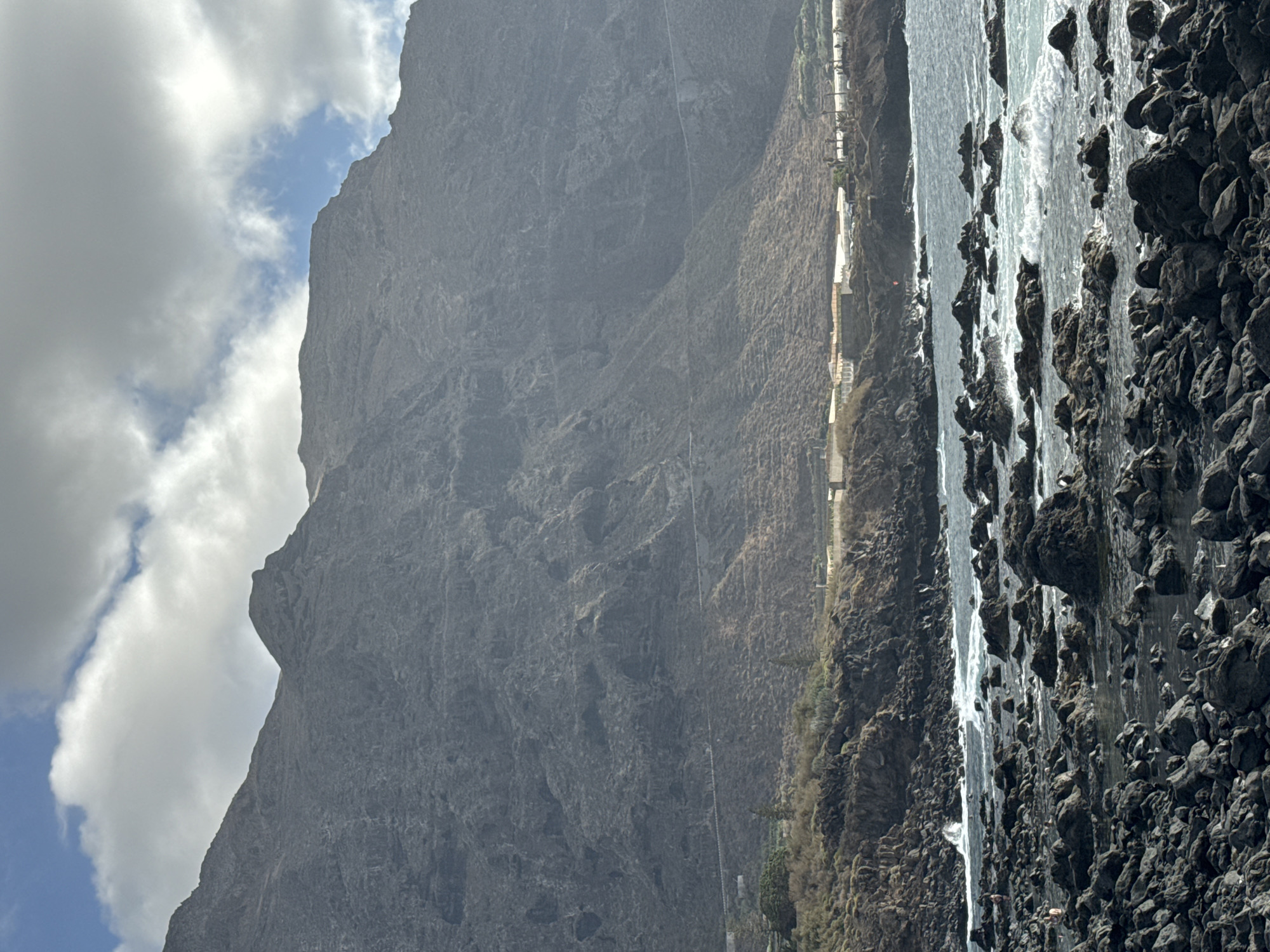 The image shows a rocky coastline with a large, rugged mountain dominating the background. The mountain is steep and appears to be covered in dry, scrubby vegetation in some areas, with evidence of a road winding along its lower slopes. In the mid-ground, there are several white structures that appear to be greenhouses or agricultural facilities, situated on a shelf below the mountain. A small number of people are visible on the rocky shoreline and in the water. The sea is a deep blue, with white waves crashing against the dark, jagged rocks that form the foreground. The sky is partially cloudy, with patches of blue sky visible between large, white clouds. The overall scene conveys a sense of natural grandeur and rugged beauty.