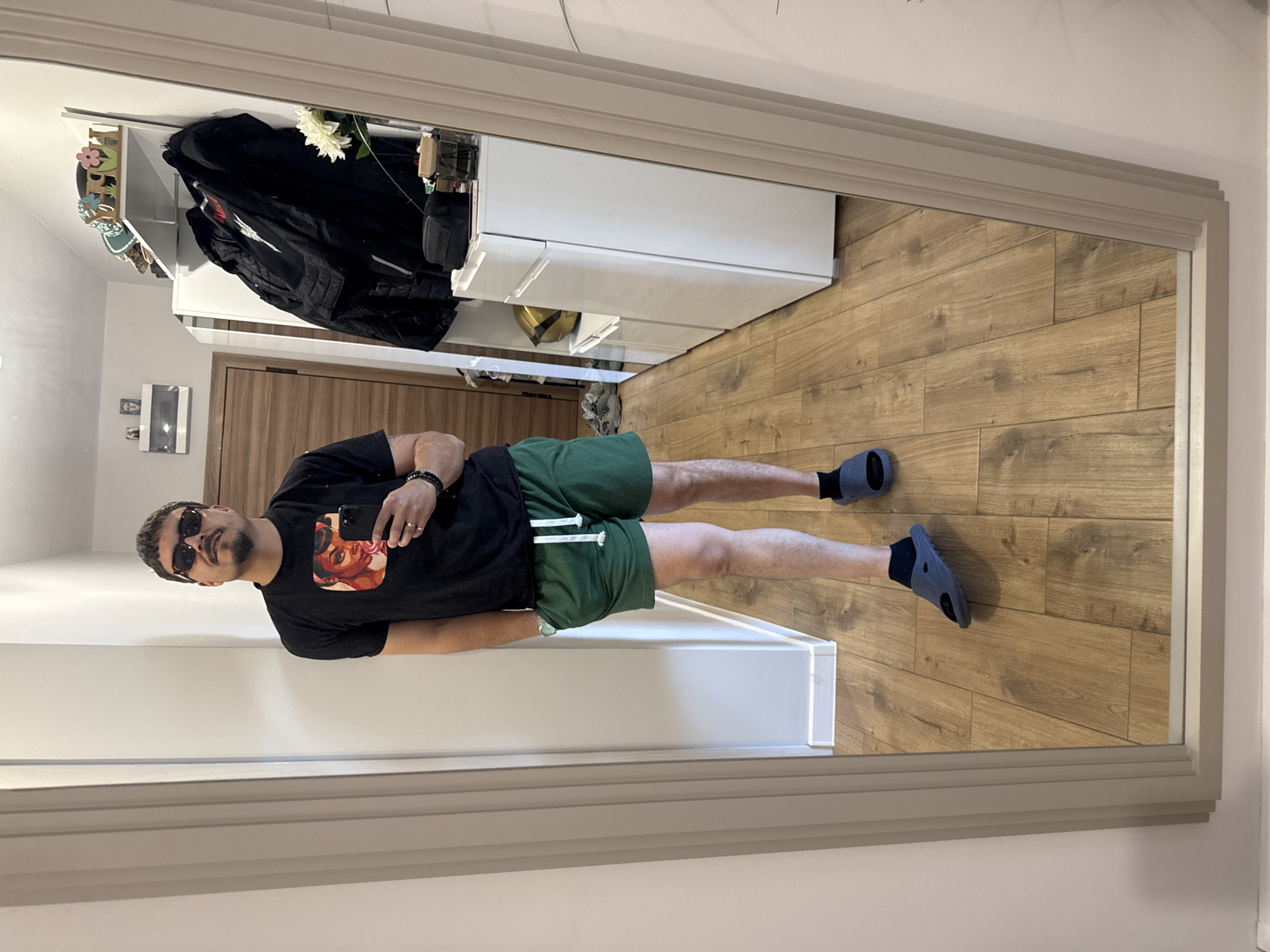 A man in sunglasses, a black t-shirt, and green shorts is reflected in a mirror. He is holding a phone in his left hand. The reflection shows a hallway with wooden floors, a white cabinet, and a dark jacket hanging inside. The location appears to be a home in an unknown city.