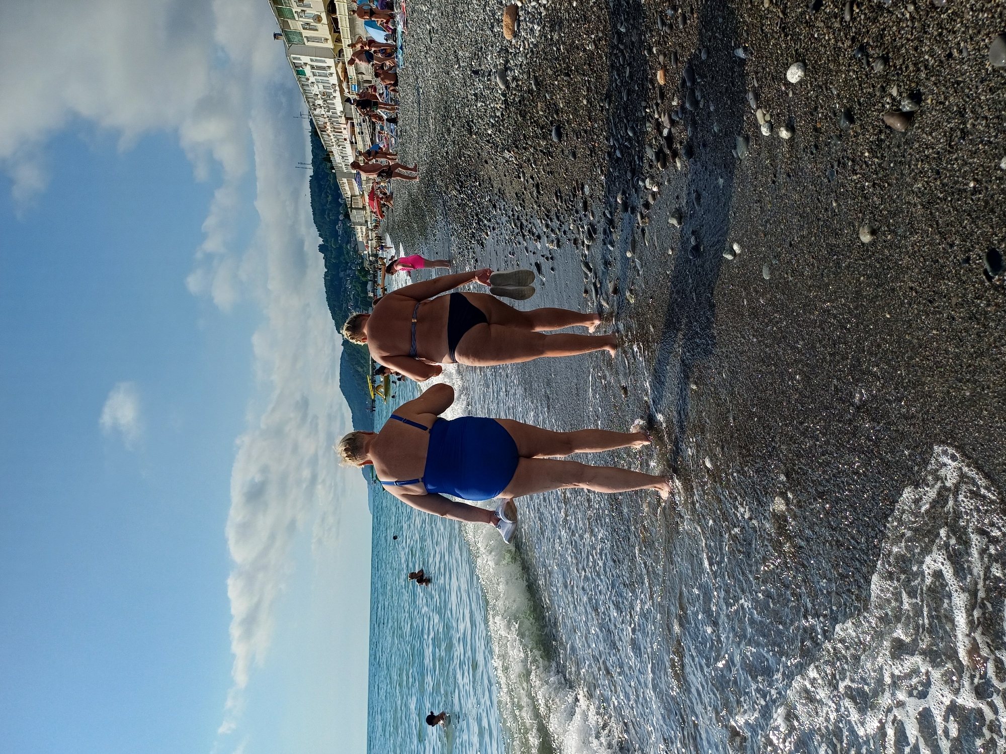 This is a full-shot, eye-level, outdoor photograph of a pebble beach in Sochi, Russia, on a sunny day with some clouds. Two adult women, appearing to be of similar age and size, are walking into the shallow water from the shore. Both are wearing swimsuits. The woman on the left is wearing a blue one-piece swimsuit and carrying a pair of white flip-flops in her right hand. The woman on the right is wearing a dark bikini and carrying a pair of light-colored flip-flops in her right hand. Gentle waves are washing onto the shore around their feet. In the background, other beachgoers are visible, some in the water and others on the shore near buildings and umbrellas. The sea extends to the horizon, where it meets a hazy sky with scattered white clouds. Mountains covered in trees are visible in the distance. The buildings along the coastline appear to be hotels or apartments, characteristic of a resort town.