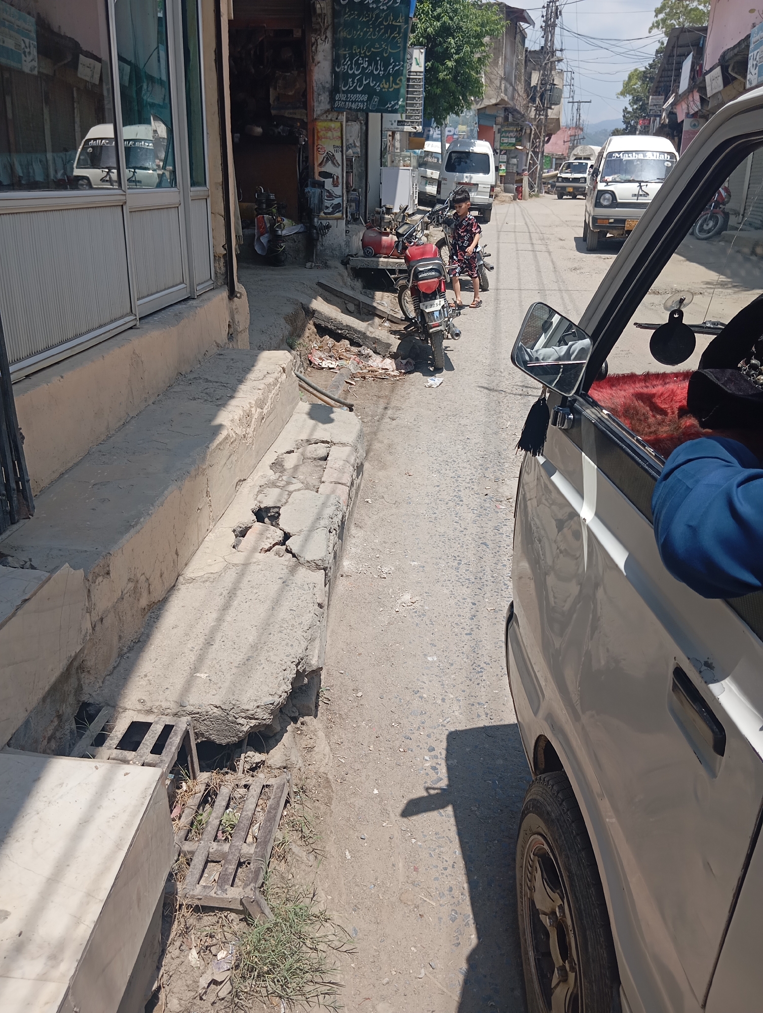 The image depicts a street scene in Mansehra, Pakistan, likely during daytime. A white Suzuki van is prominently featured, with its passenger side visible from a low angle, suggesting the photo was taken from inside another vehicle. The van's interior shows a red fuzzy seat cover and a person's arm wearing a blue sleeve. The exterior of the van is light-colored and shows some minor scuffs.
The street itself is paved with asphalt and shows signs of wear and tear. To the left of the van, a raised concrete sidewalk or platform is visible, with sections that appear cracked and broken. A metal grate covering a drain is also present on this platform, with some grass growing around it.
On the street, several motorcycles are parked along the left side. One motorcycle, a red and silver model, is particularly visible. A young boy, wearing a dark patterned shirt and shorts, stands near this motorcycle. Further down the street, other vehicles, including white vans and motorcycles, are visible, indicating typical traffic in a Pakistani city. Buildings line both sides of the street, with storefronts displaying various signs, some of which appear to be in Urdu. Trees and utility poles are also present, contributing to the urban environment. The overall impression is one of a busy, functional street in a Pakistani town.