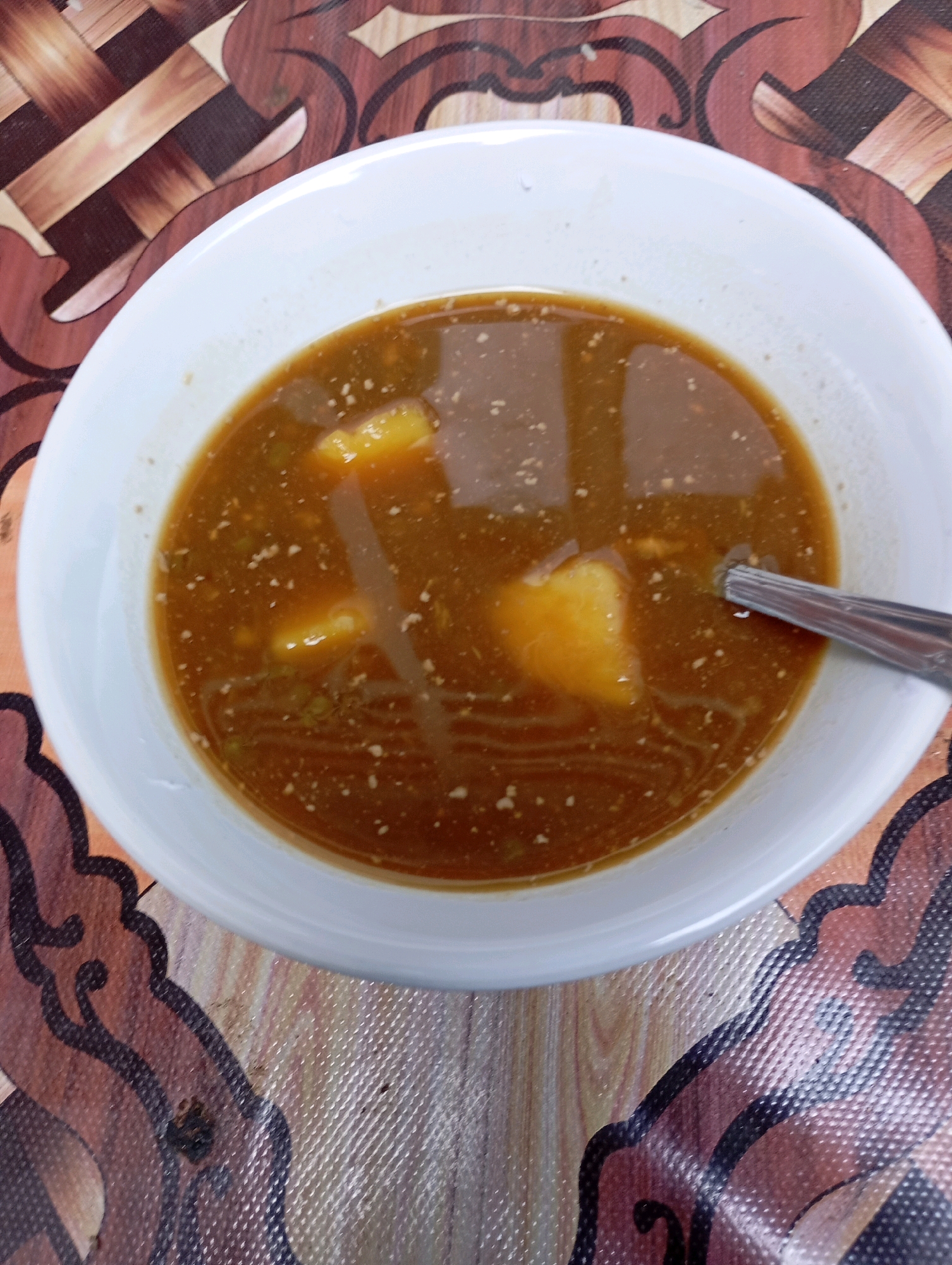 A white bowl containing a brownish liquid with chunks of potato and what appears to be minced meat is placed on a patterned placemat with brown and tan wavy designs. A silver spoon rests in the bowl. The scene appears to be from a domestic setting in Balikpapan, Indonesia.