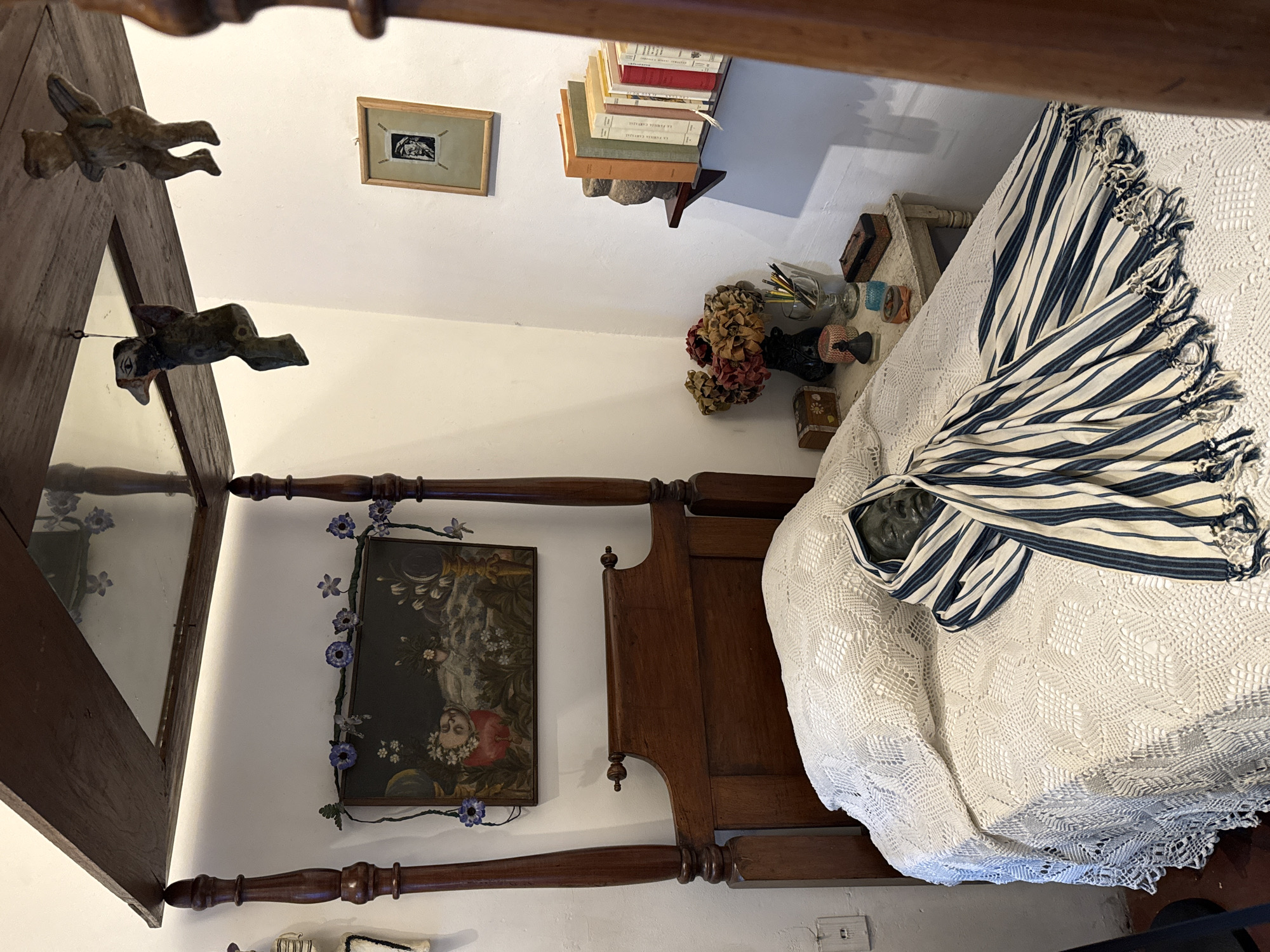 The image shows a bedroom in the Casa Azul (Blue House), the childhood home and later residence of Frida Kahlo, located in Coyoacán, Mexico City, Mexico. The room contains a four-poster bed with a white crocheted bedspread. A blue and white striped shawl is draped over the head of a sculpted head lying on the bed. A painting of a woman surrounded by flowers hangs on the wall adjacent to the bed. A mirror with wooden frame adorned with winged animal figures is visible. A small framed picture hangs above a bookshelf filled with books. A vase of dried flowers and other items are positioned on a shelf next to the bed. The room has white walls.