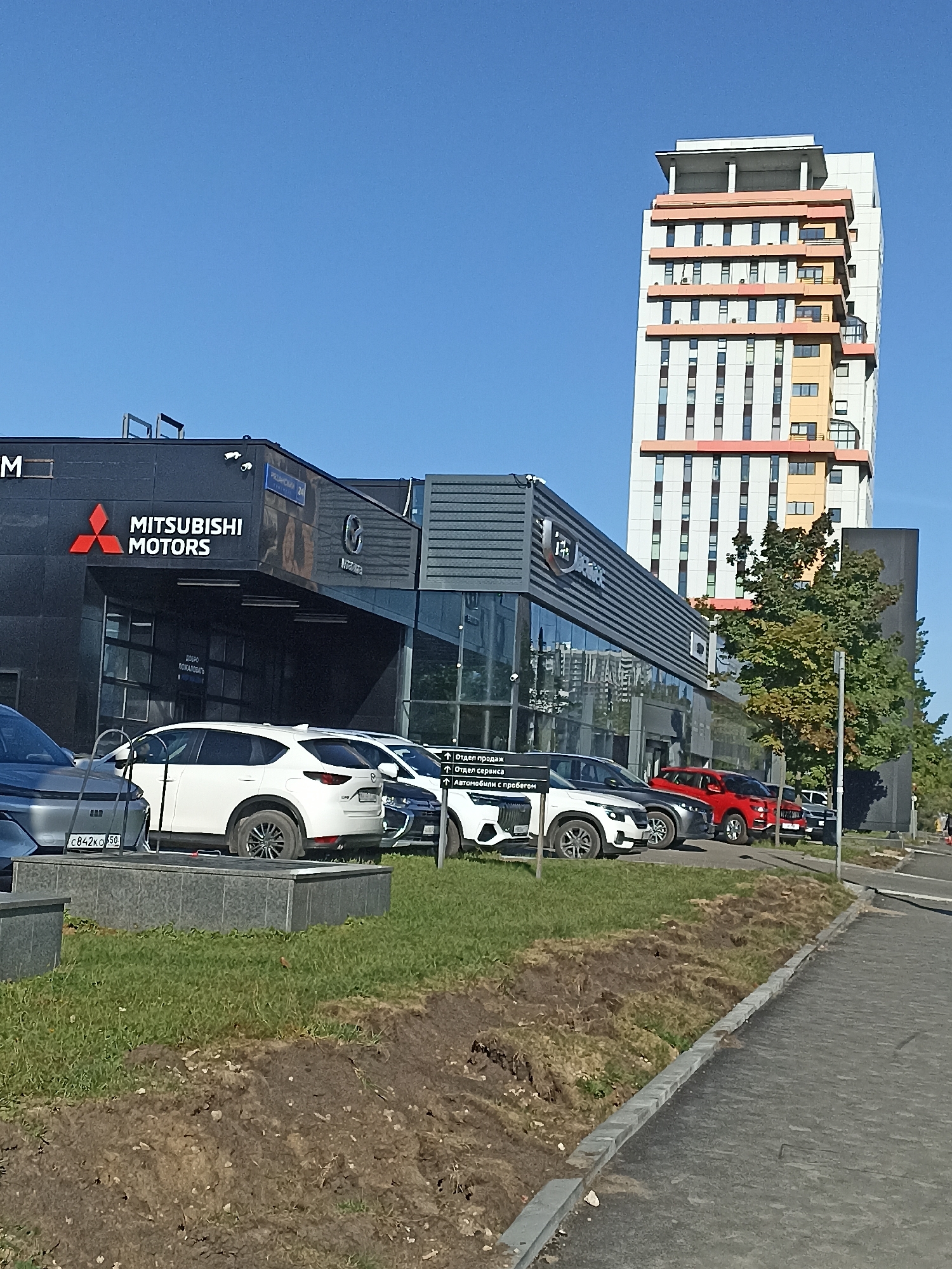 The image shows a car dealership with a Mitsubishi Motors sign. The dealership is located in Moscow, Russia, as indicated by the license plate on the gray car in the foreground. The dealership building is modern with large glass windows. Several cars are parked in front of the dealership, including a white SUV, a red car, and a gray car. In the background, a tall, modern apartment building stands against a clear blue sky. The scene is well-lit, suggesting it is daytime. There are no people clearly visible in the image. The ground in front of the dealership is covered with grass and some bare soil, indicating recent landscaping or construction. A paved walkway runs along the right side of the image.