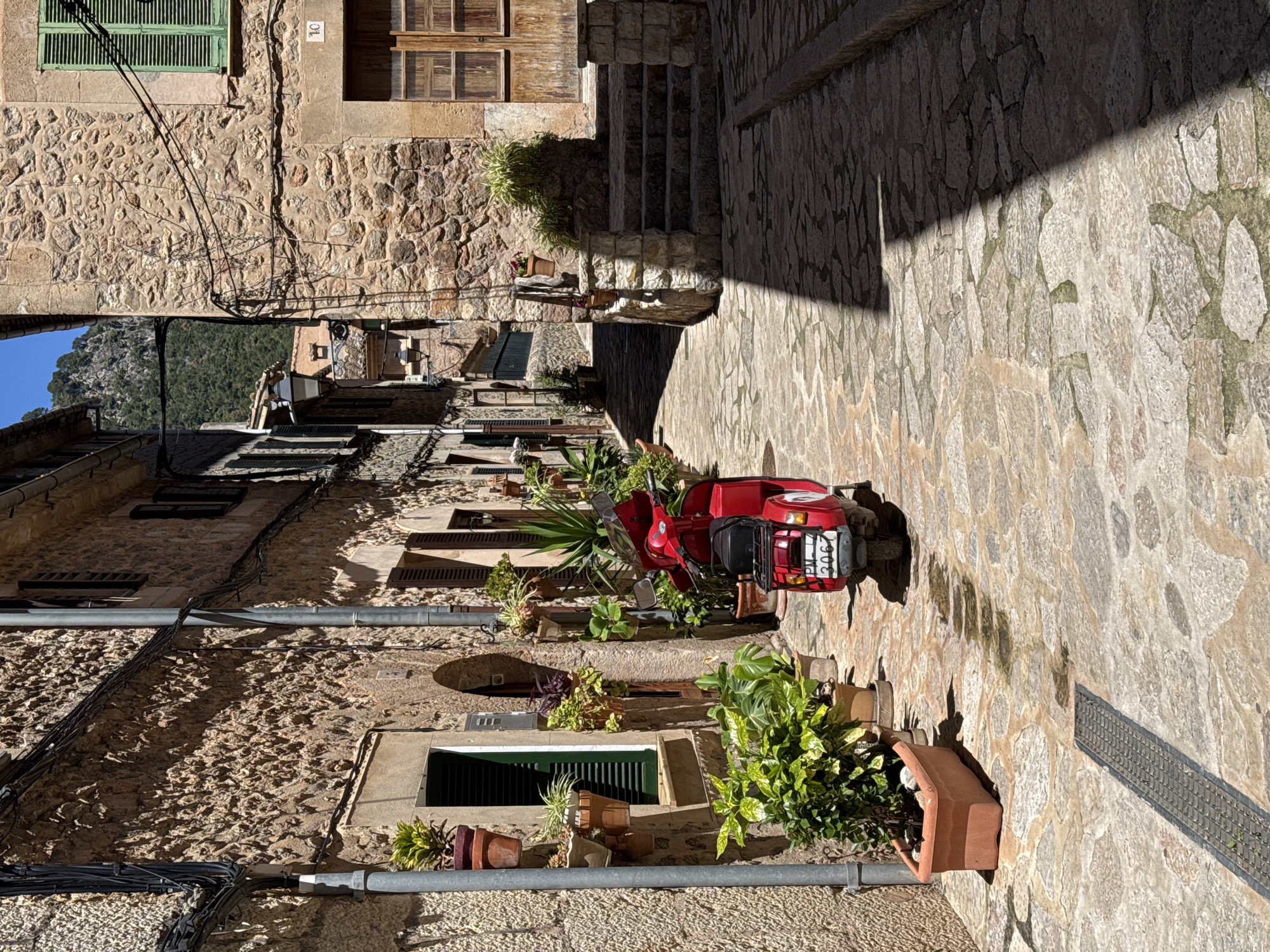 The image shows a narrow street in Valldemossa, Spain, lined with stone buildings. A red scooter is parked on the cobblestone street. Various plants in pots are placed along the building walls. A drainage grate runs along the edge of the street. A building with a green shuttered window is visible. In the distance, the buildings create a tunnel view, with mountains visible at the end. The image appears to be rotated 90 degrees clockwise.