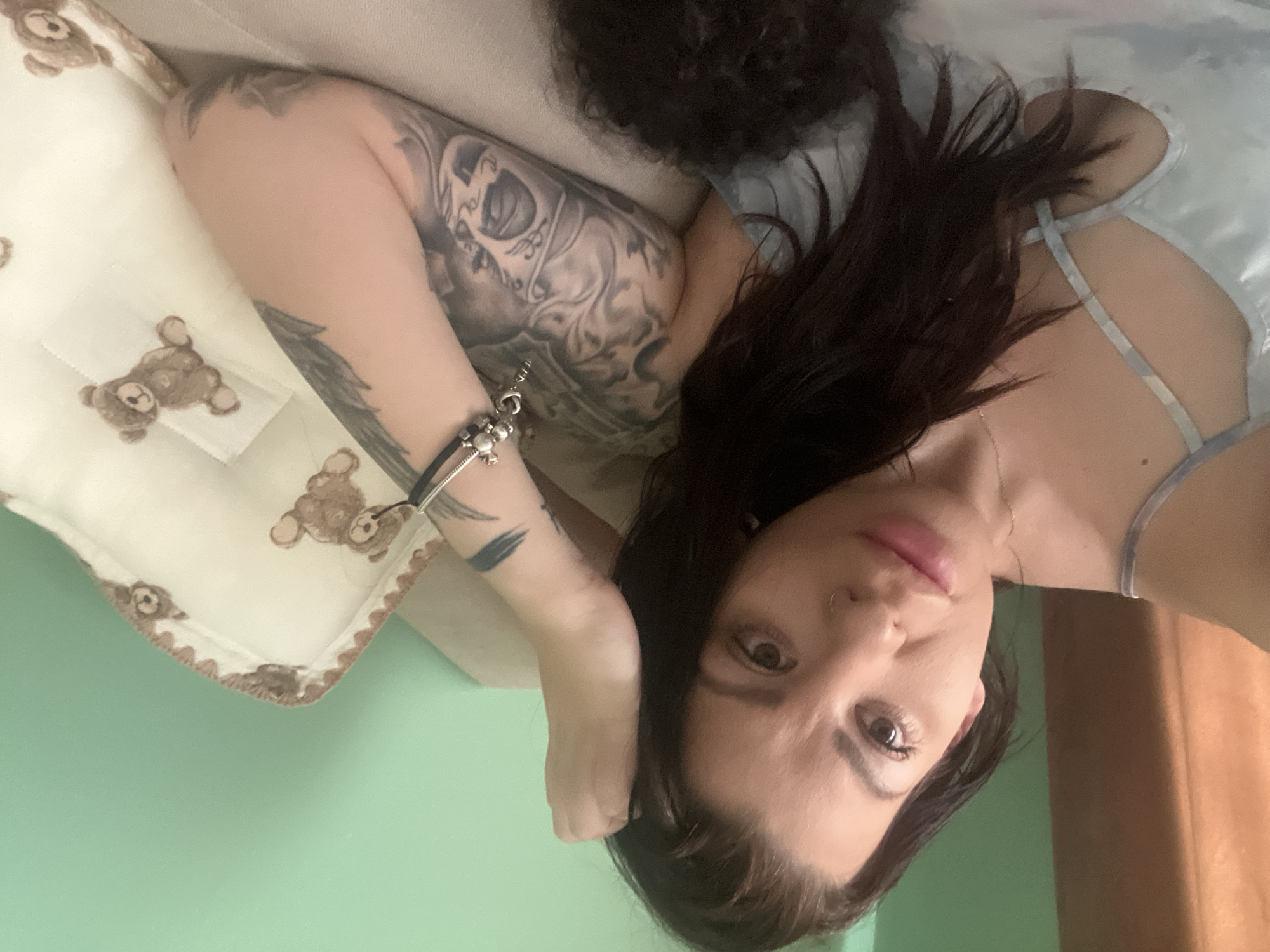 A woman with dark hair and visible tattoos on her arm is lying down, propped up on her elbow. She is wearing a light-colored camisole. Her hand rests on her head, and her arm is adorned with a bracelet. Behind her, a pillow with a teddy bear pattern is visible against a teal-colored wall. To the left, a brown curtain is partially visible. The overall setting suggests an indoor residential environment, possibly a bedroom or living area. The image quality is that of a casual selfie. There is no information in the image to suggest a specific location in Plovdiv, Bulgaria.