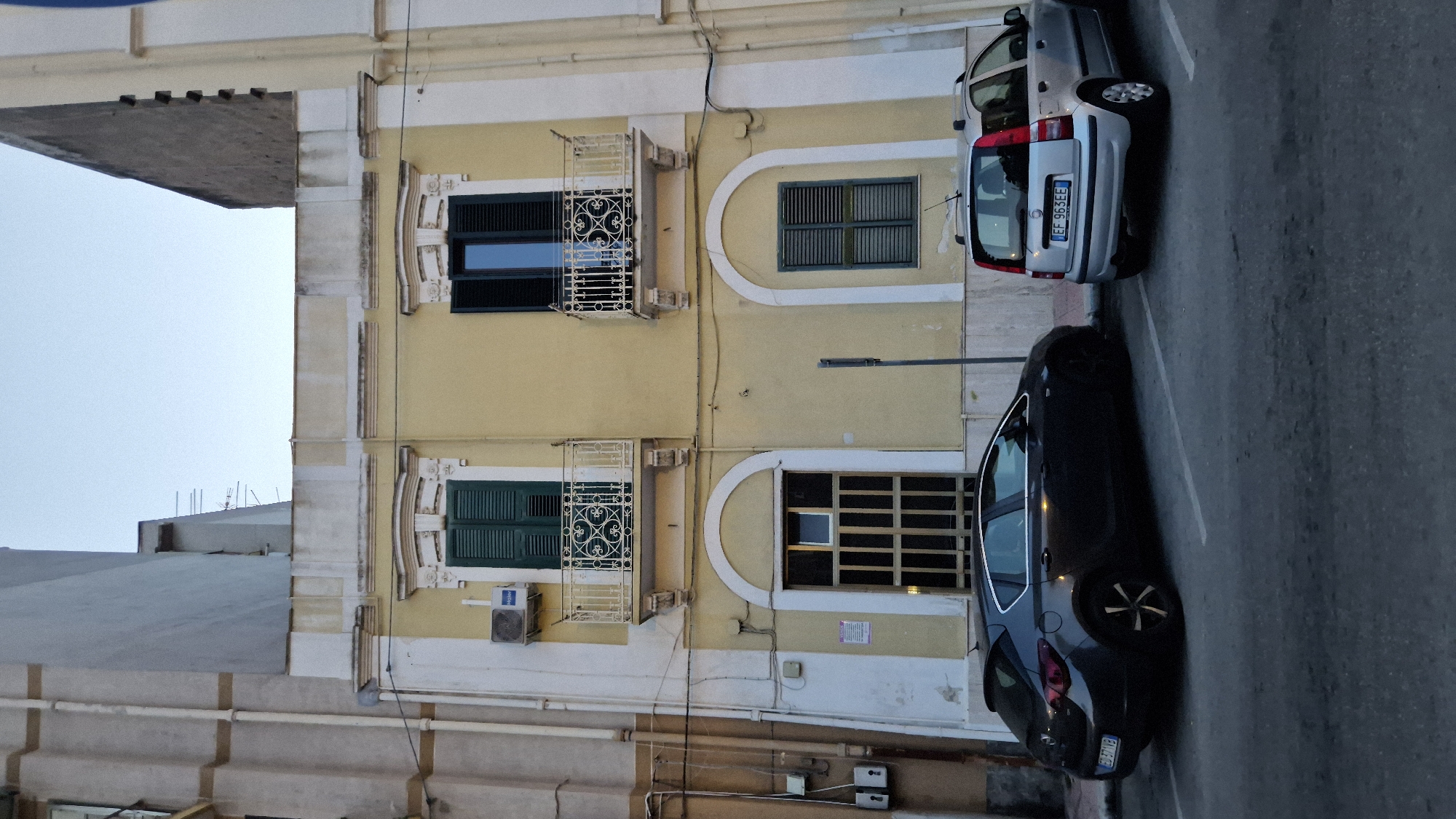 The image shows a street scene in Taranto, Italy, with a pale yellow building prominently featured. The building has two stories with arched windows on the ground floor and rectangular windows on the upper floor, each with green shutters and decorative wrought-iron balconies. An air conditioning unit is visible on the left side of the upper floor. Two cars are parked in front of the building: a dark grey hatchback on the left and a silver compact SUV on the right. The street surface is asphalt, marked with parking lines. The sky is overcast.