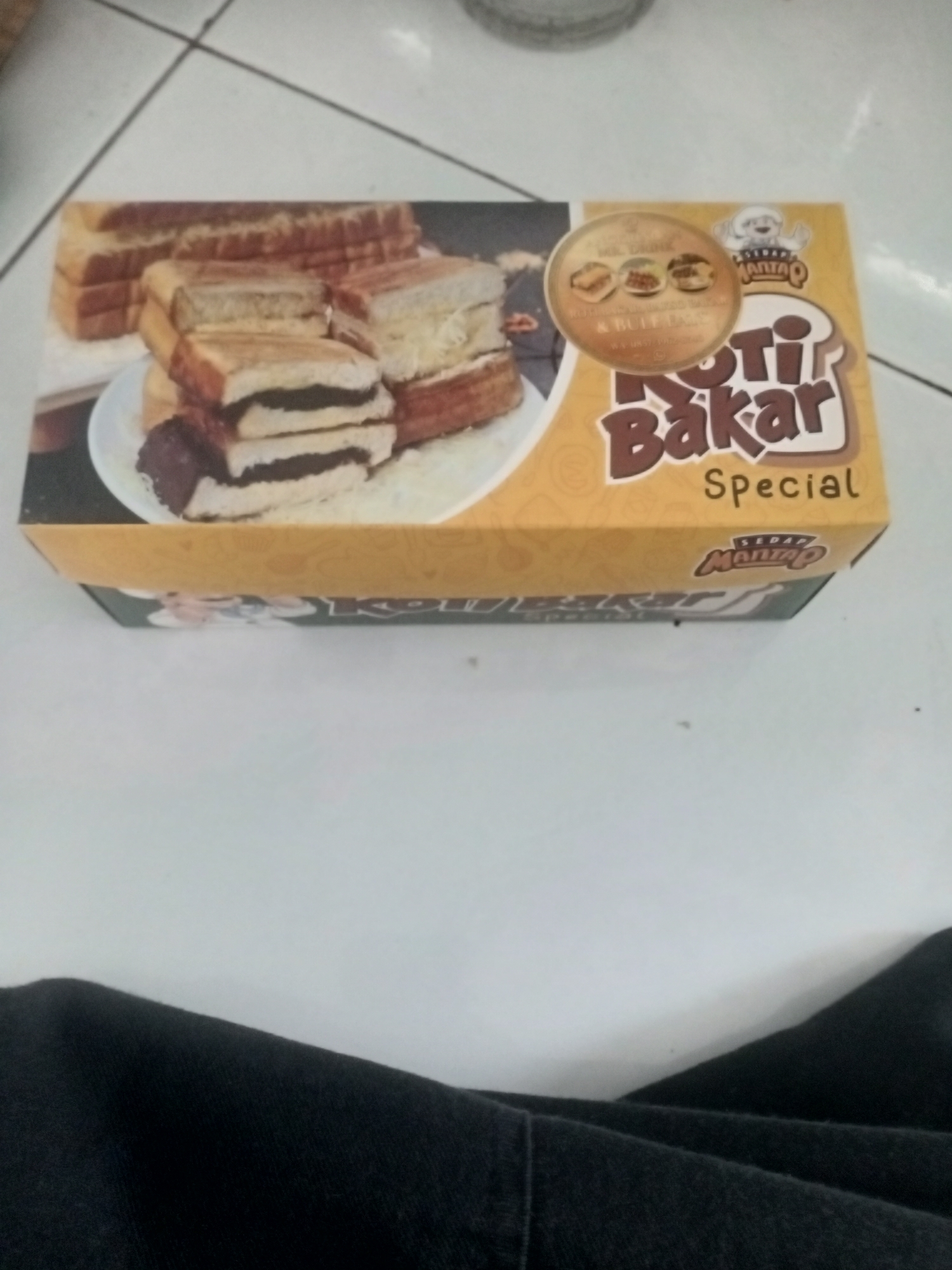 A box of "Roti Bakar Special" (Special Toasted Bread) is presented on a tiled floor, likely within a commercial establishment in Badas, Indonesia. The box features an illustration of several slices of toasted bread, some filled with dark chocolate and others with white filling, suggesting various flavor options. The "Roti Bakar" branding is prominent, with a stylized loaf of bread and a mascot of a person in a chef's hat. Below this, "Special" is written in smaller text. The box itself is yellow with brown accents. The tiled floor is light-colored with dark grout lines, and a portion of black fabric, possibly clothing, occupies the bottom of the frame.