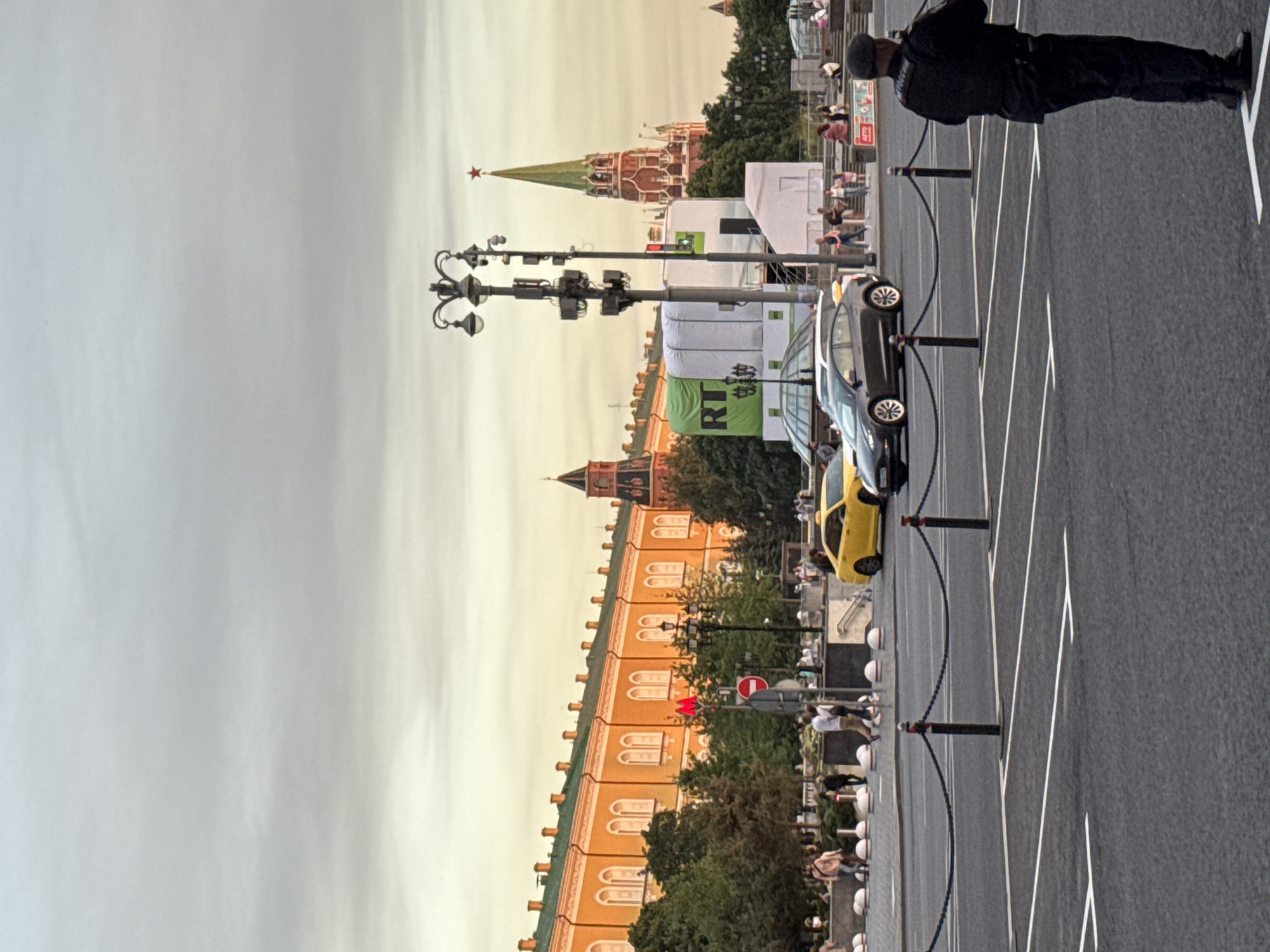 The image captures a street scene in Moscow, Russia, near the Kremlin. In the foreground, a person in a dark uniform, possibly a security guard or police officer, stands facing away from the camera, looking towards the left of the frame. The ground is paved with asphalt, marked with white lines indicating parking spaces. Black bollards connected by chains delineate the parking area from the street.
In the middle ground, a grey car is parked on the left, and a yellow taxi is visible on the right. Behind them, a street with pedestrians walking on a sidewalk is discernible. Several trees provide greenery along the street. A lamppost with multiple lights stands prominently. To the left of the lamppost, there's a building with an orange facade, featuring arched windows. A red "M" sign and a circular red and white sign are also visible.
In the background, iconic Moscow landmarks are present. A tall, green-roofed tower with a red star on top, characteristic of the Kremlin, dominates the right side of the frame. Adjacent to it, another red tower with similar architectural features can be seen. A large white structure with a green banner displaying "RT" is positioned in front of these buildings. The sky is overcast, with a diffused light suggesting it might be late afternoon or early evening. The overall scene depicts a public space with a mix of modern vehicles, historical architecture, and people in an urban environment.