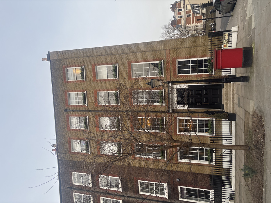 The image displays a multi-story brick building from a street-level perspective, with the original image rotated 90 degrees clockwise. When viewed correctly, the building stands on the left side of a paved sidewalk, with a street visible further to the right.
The building itself is tall and narrow, featuring a facade primarily composed of light brown or yellowish bricks with red brick detailing around the windows and as corner quoins. It appears to be at least four or five stories high. Each story has multiple rectangular windows with white frames. Many of the windows on the upper floors contain decorative green foliage, possibly window boxes or wreaths, giving a festive or well-maintained appearance. Some windows show interior lighting, suggesting it might be late afternoon or evening, or simply that lights are on inside.
At the ground level, there is a prominent black double door serving as the main entrance, flanked by large windows. A traditional black street lamp stands directly in front of the building, to the right of the entrance. A black metal fence or railing runs along the base of the building, separating it from the sidewalk.
To the right of the building, on the sidewalk, a bright red cylindrical pillar box (postbox) is clearly visible. Attached to the postbox or a nearby pole is a white street sign with black lettering that reads "BEDFORD ROW" and below it, "WC1". Further down the street, an older, more ornate brick building with gables and multiple windows can be seen in the background, suggesting a historical urban setting. Cars are parked or driving on the street beyond the sidewalk.
A bare-branched tree, possibly deciduous and in winter, stands in the foreground, its branches partially obscuring the front of the building. The sky above is overcast and light gray, indicating a cloudy day. The overall impression is one of a classic London street scene.