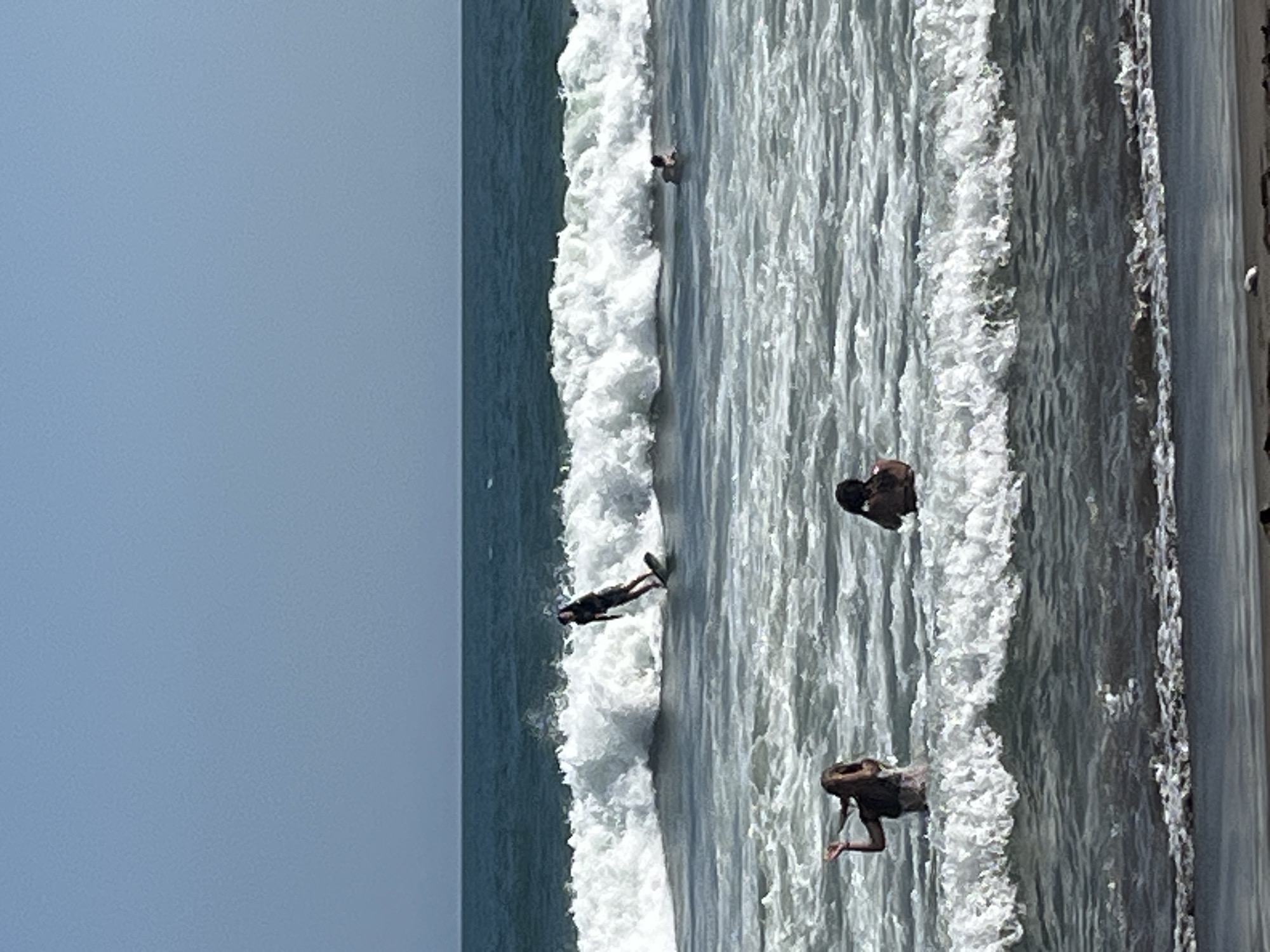 The image depicts a scene at a beach, likely in North Kuta, Indonesia, with a clear blue sky and a choppy ocean. In the foreground, two individuals are visible in the water, with one in the process of standing up and another seated. A large wave is breaking in the middle ground, with a surfer riding its crest. Further in the distance, another individual is visible in the water. The shoreline is visible at the bottom of the image, with a small object that could be a rock or shell on the sand.