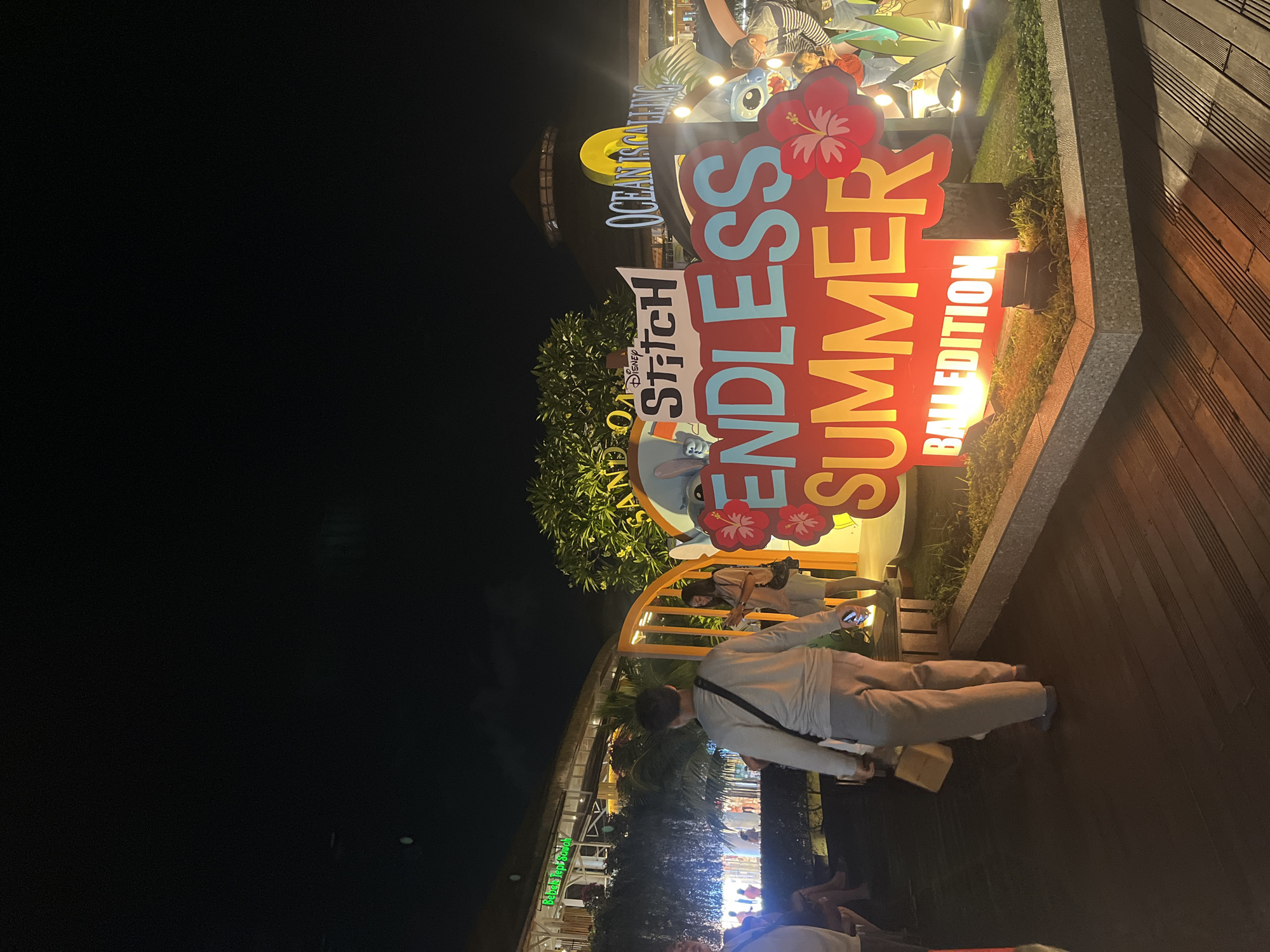 The image depicts an outdoor scene at night, possibly a plaza or walkway, in Kuta, Indonesia. A prominent signage displays "ENDLESS SUMMER BALL EDITION" in large, illuminated letters, with "Disney Stitch" and "OCEAN IS CALLING" also visible. Several people are present, including a man walking away from the camera, a woman standing near the signage, and a few figures in the background. The environment features wooden decking, decorative greenery, and architectural elements of buildings in the background, illuminated by artificial lights. The overall atmosphere is festive and commercial.
