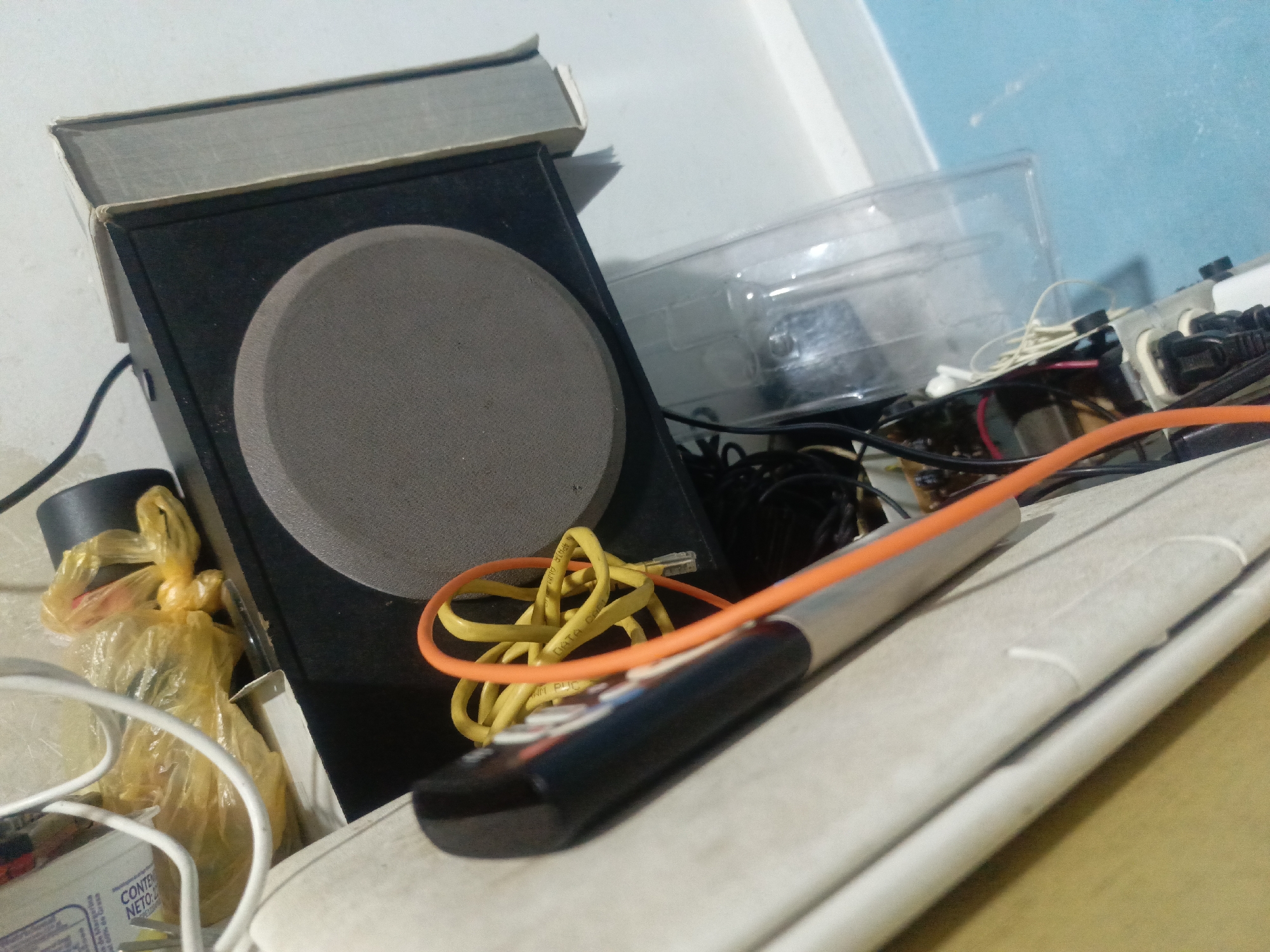 The scene appears to be a cluttered desk or shelf in an indoor setting. A black speaker with a grey circular cone is present, topped by a book. In front of the speaker, a remote control rests atop a white object (possibly a printer or another device). Tangled yellow and orange cables are intertwined around the remote and speaker. A clear plastic container is visible towards the upper right, along with an assortment of wires and electronic components. There are glimpses of packaging and plastic bags. No people are visible. The location, Juan Griego, Venezuela, cannot be confirmed from the image alone.