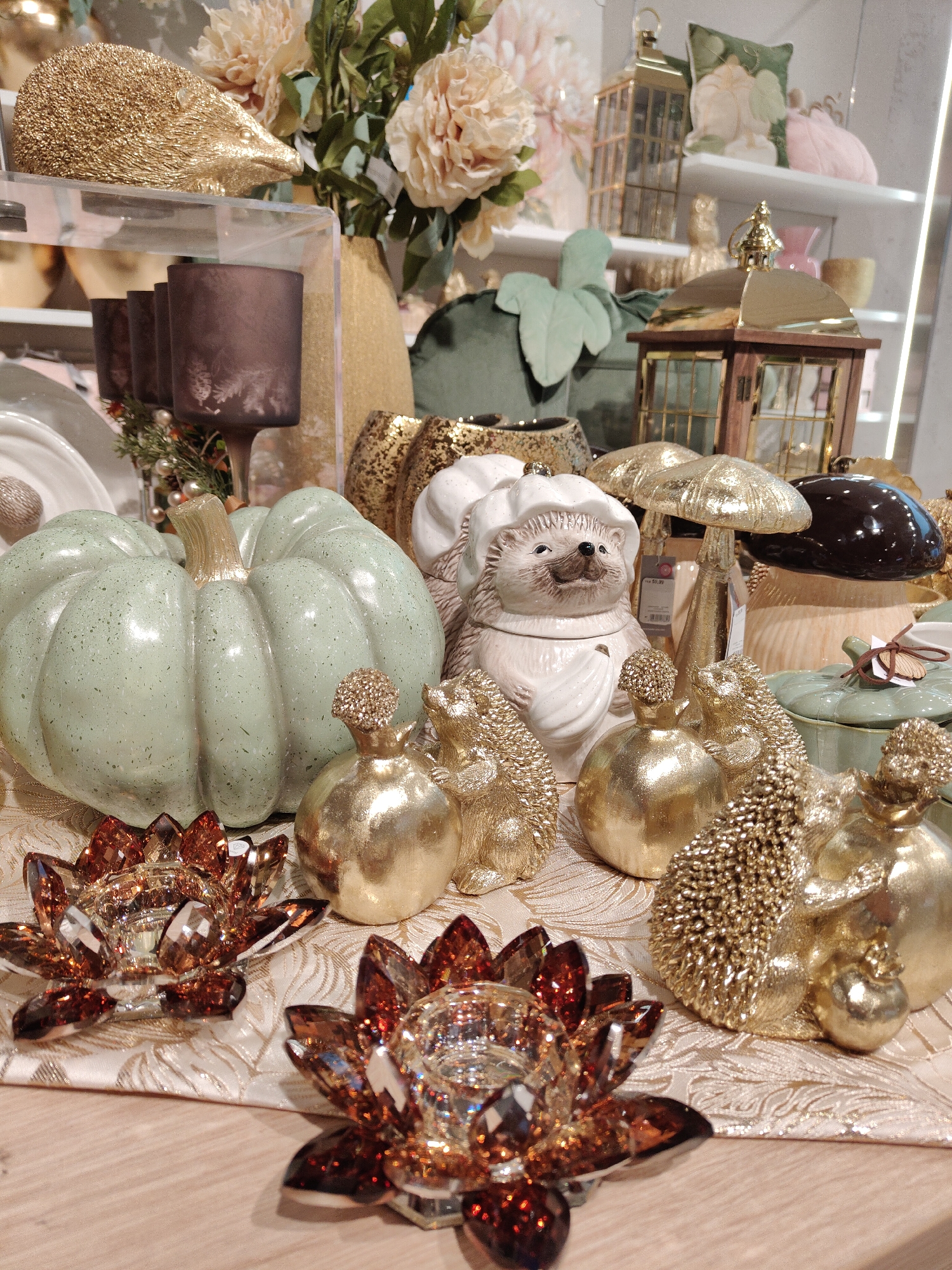 The image shows a tabletop display of decorative autumn-themed items, likely in a store or home. In the foreground, there are two lotus-shaped candle holders made of amber-colored faceted glass. Behind them, the table is covered with a beige cloth featuring a subtle floral pattern.
The main subjects are various decorative figurines. A large, pale green ceramic pumpkin with a gold stem is prominently placed. To its right, a white ceramic hedgehog figurine wearing a chef's hat and apron is visible, appearing to hold a small spoon or whisk. Several small, gold hedgehog figurines are also present, along with gold pomegranate-shaped decorations. Further back, there are gold mushroom figurines and a dark brown, glossy mushroom-shaped object.
In the background, there are more decorative elements, including tall glass vases with dark purple candle holders, a golden lantern, and a stack of shelves displaying more decorative items such as floral arrangements and possibly more pumpkins. The overall impression is one of seasonal decoration, with a focus on autumn colors and textures like gold, green, and amber.
Without additional context or visual cues, it is impossible to determine the specific location within the city of Gdynia, Poland, or to identify any actions or interactions of people. The image focuses solely on the static arrangement of decorative objects.