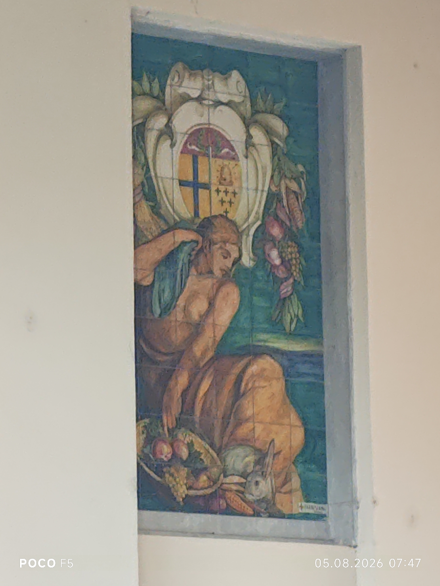 This is a photograph of a colorful ceramic tile mural depicting a classical scene. The mural is framed by a light-colored border and is set into a wall. The mural itself is vertically oriented and features a coat of arms at the top, adorned with scrolls and foliage. Below the coat of arms, a reclining female figure is depicted, draped in fabric. She is holding a piece of cloth and appears to be looking downwards. In front of her, a basket is filled with fruits such as apples and grapes, and a rabbit is nestled among the produce. The background of the mural is a deep teal color. A faint signature is visible at the bottom right of the mural. The overall style of the artwork is reminiscent of Renaissance or Baroque art. The photograph also includes a timestamp in the lower right corner: "05.08.2026 07:47". The POCO F5 logo is present in the lower left corner. This mural is located on the facade of the "Piscina Comunale" (Municipal Swimming Pool) in Collecchio, Italy.