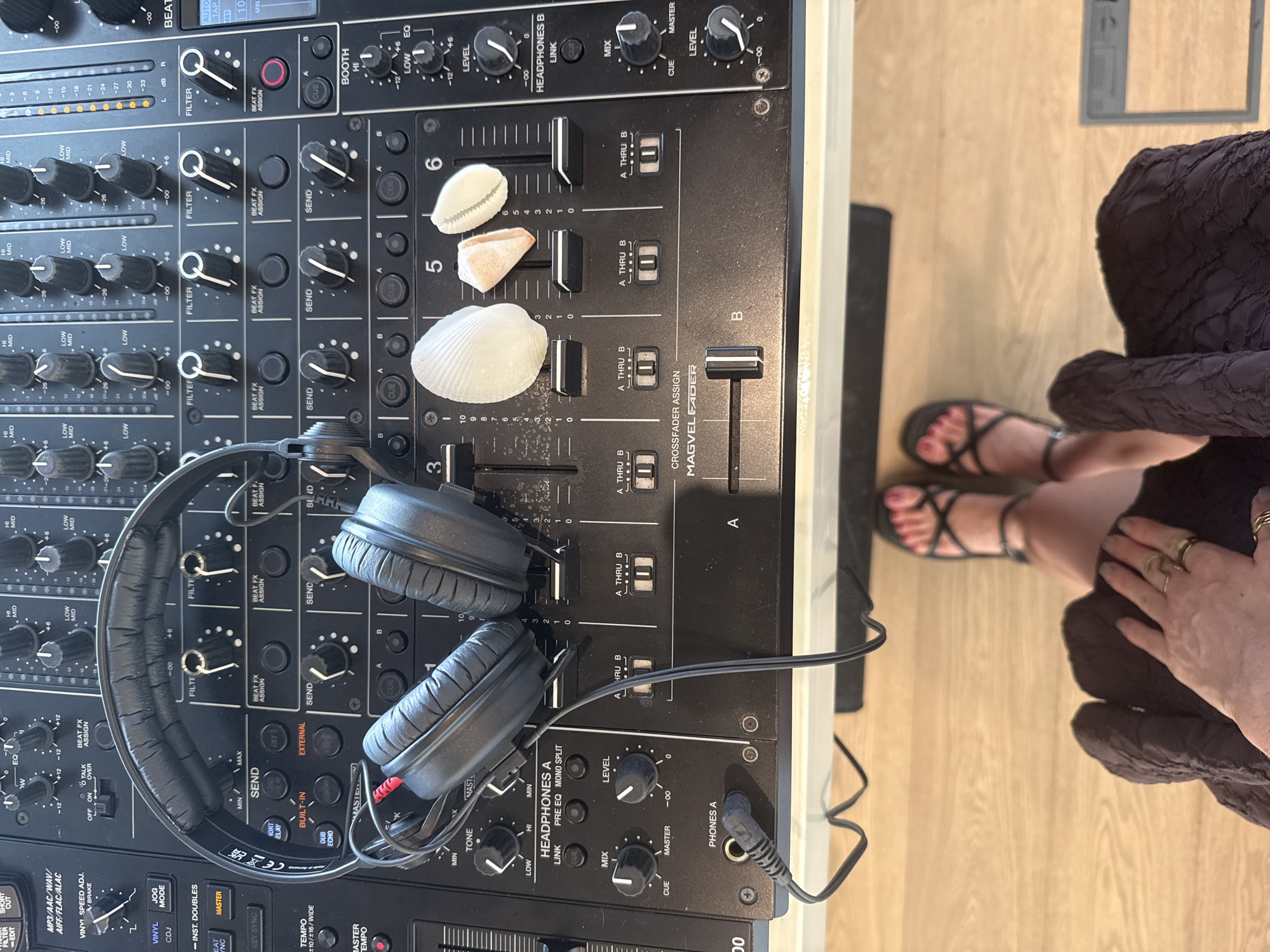 The image shows a close-up, overhead view of a DJ mixer with headphones placed on top. The mixer has numerous knobs, faders, and buttons, consistent with professional audio equipment. Three seashells are placed on the mixer surface. In the lower part of the image, a person's legs and feet, adorned with sandals, are visible, along with their hand resting on their lap. The background appears to be a wooden floor. The scene suggests a casual, possibly domestic, setting where someone is engaged with DJ equipment. The provided location of "Unknown, Maldives" does not correlate with any visible elements in the image, as there are no specific environmental or architectural cues to suggest this location.