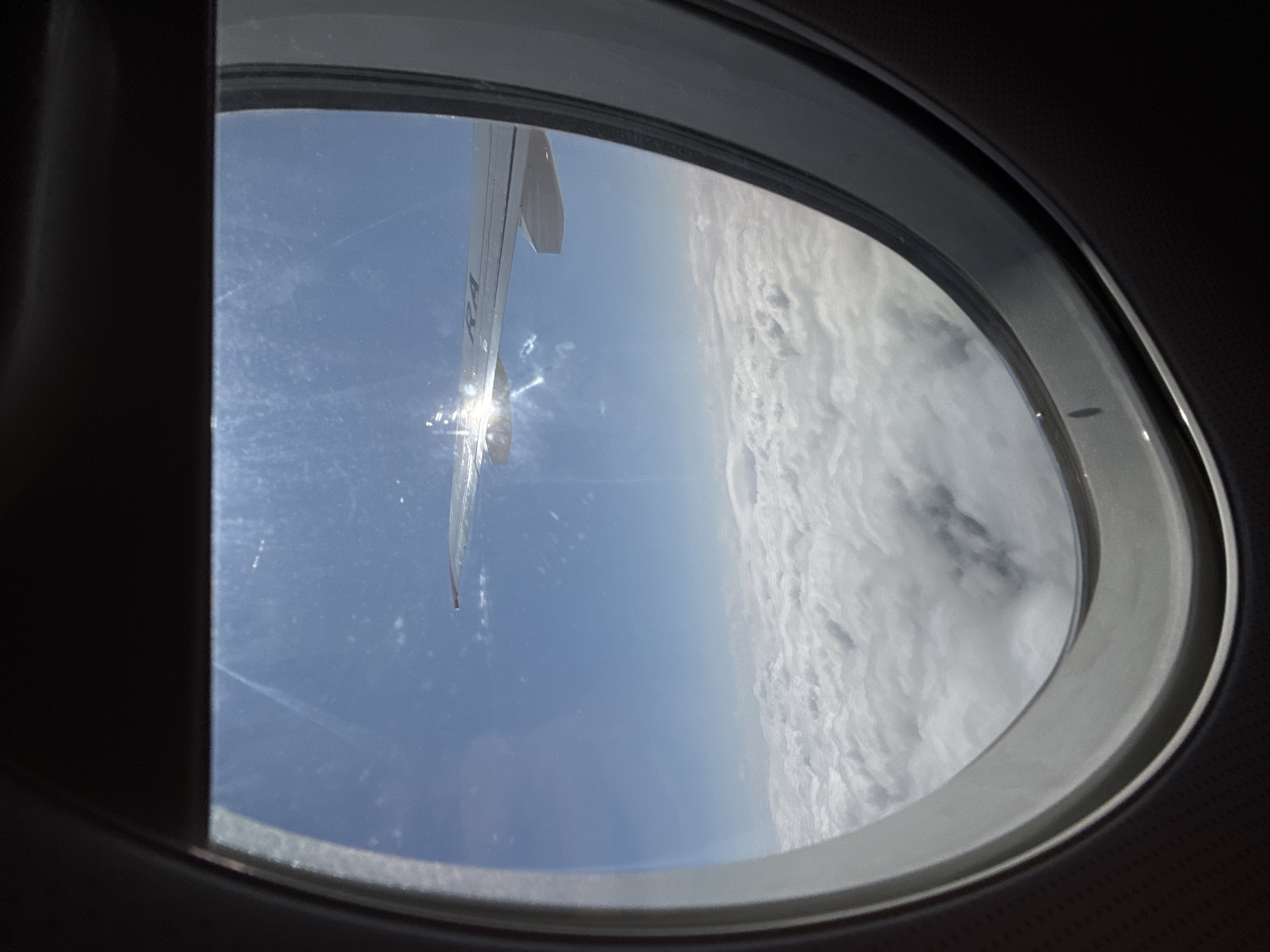 The image is taken from inside an airplane window, looking out at a vast expanse of white clouds beneath a bright blue sky. The sun is shining intensely, creating a strong lens flare on the window and casting a glint on the airplane's wing, which is visible extending into the frame from the left. The wing appears to have some faint markings on it, including what looks like the letters "RA" in an angular font. The window frame, dark and textured on the left and lighter and smoother on the right, surrounds the view. The scene depicts typical conditions during an airplane flight above the clouds. The location is identified as Staroe Mart'yanovo, Russia.