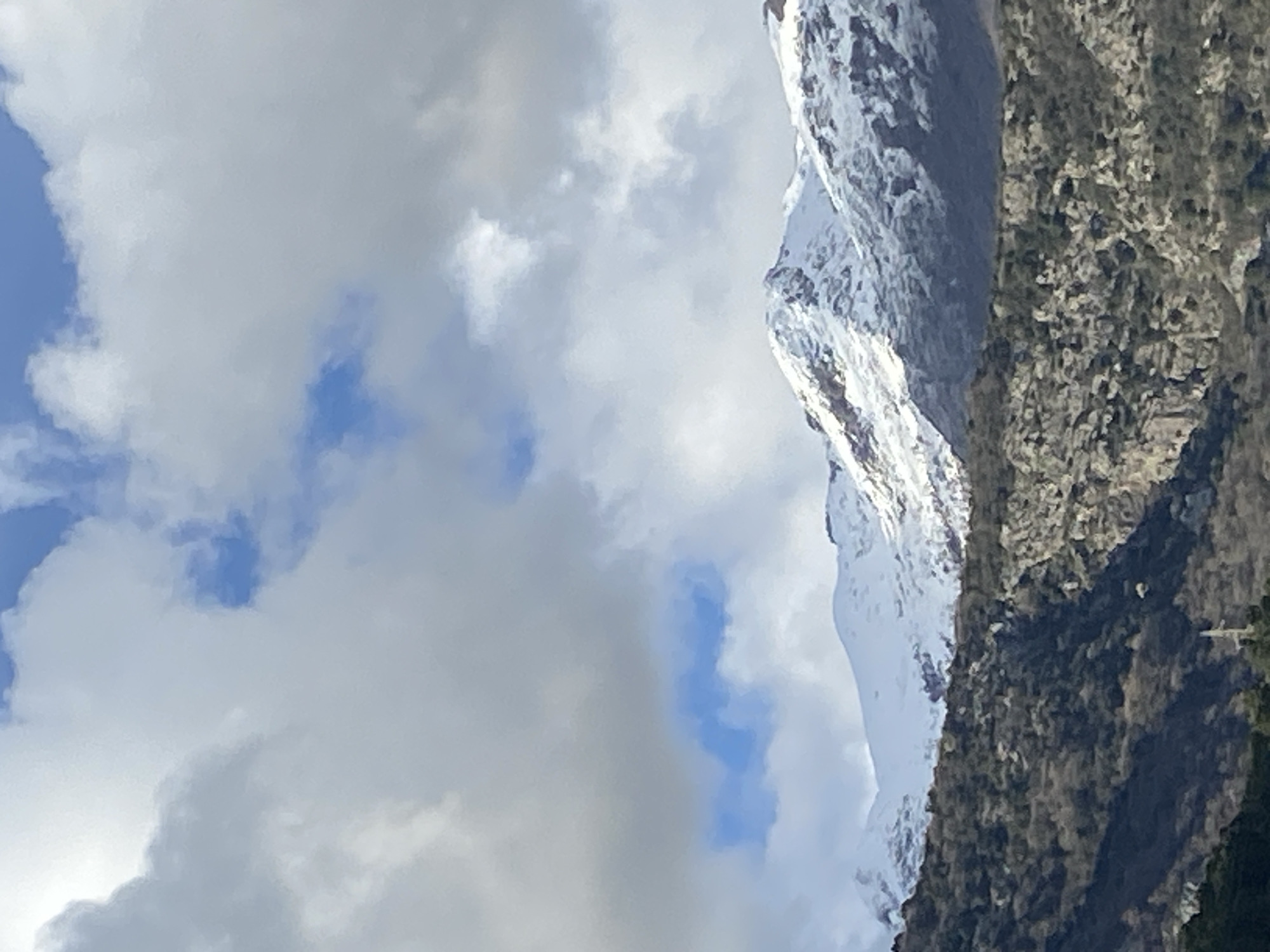 The image displays a panoramic view of a snow-capped mountain range, as observed from Budva, Montenegro. The upper 40% of the frame is dominated by a sky featuring large, light grey and white cumulus clouds, with several distinct patches of bright blue sky visible between them. Below the cloud line, a prominent mountain range extends horizontally across the middle ground, its peaks and upper slopes extensively covered in white snow, revealing darker rock faces in less covered areas. The mid-ground and foreground consist of lower mountain slopes and hills, densely covered with dark green and brown vegetation and interspersed with exposed light grey rock formations. In the lower-center, a slender, vertical light-colored mast, consistent with a communication tower or antenna, is partially visible amidst the foreground vegetation. No individuals, animals, or direct human activity are depicted in the scene.