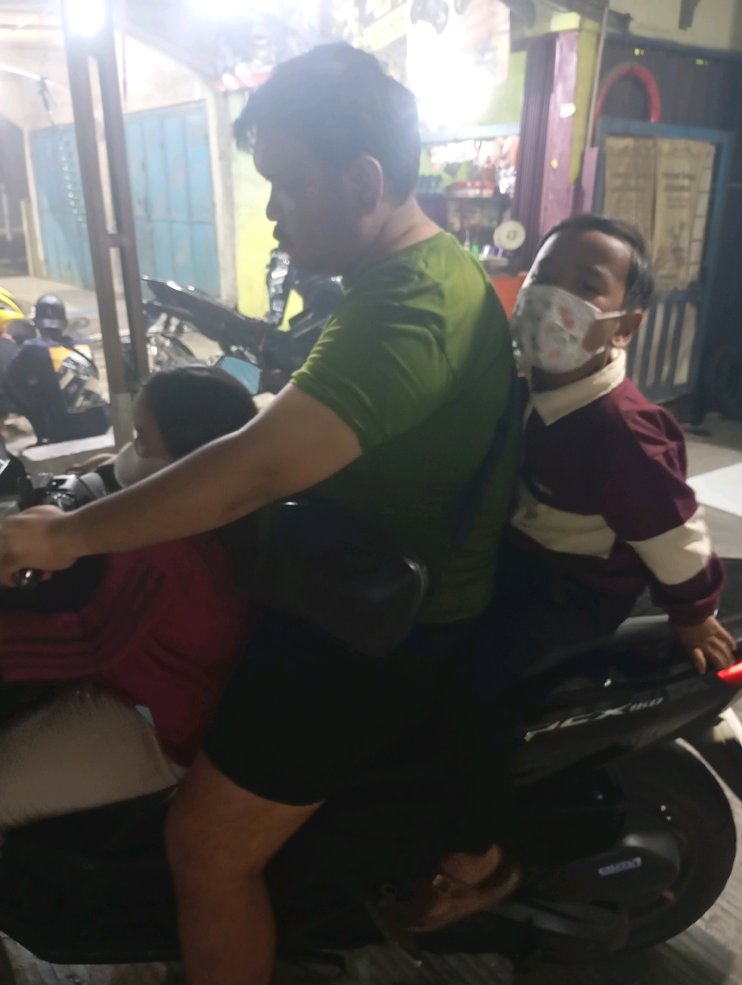 In a dimly lit urban environment, likely in Babelan, Indonesia, three individuals are seen on a motorcycle. An adult male wearing a green t-shirt is seated in the middle, operating the motorcycle. To his left, a younger individual in a red shirt, also wearing a mask, is seated behind him. To the adult's right, a child wearing a maroon and cream collared shirt and a patterned mask is seated behind the adult. The background shows a building with blue doors and some other structures, suggesting a residential or commercial area at night. The motorcycle itself appears to be a scooter-style vehicle. The overall scene captures a common mode of transportation in the region.