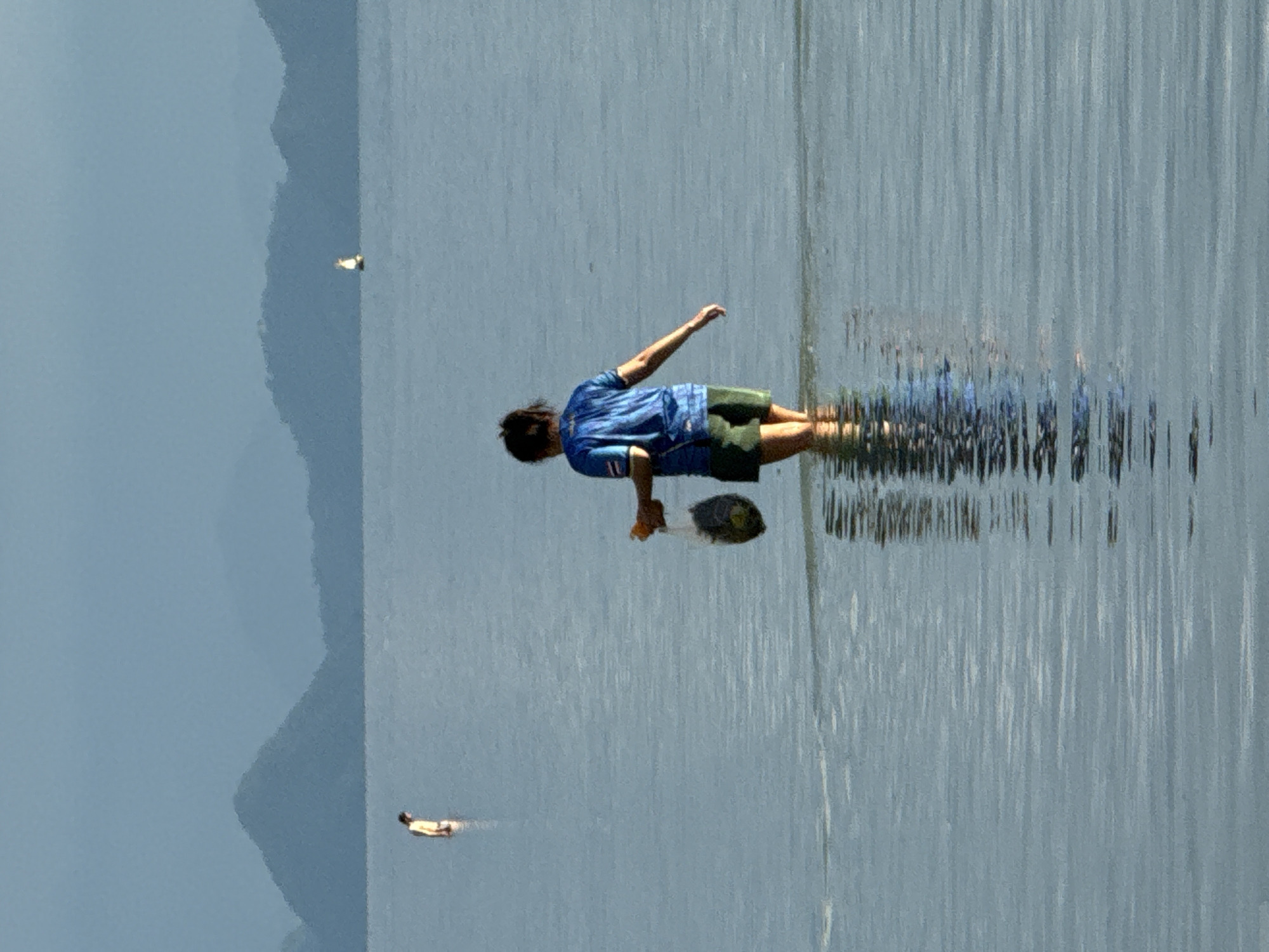 The image depicts a serene coastal scene at Baan Pang Ka, Thailand. In the foreground, a person stands in shallow water, facing away from the camera. They are wearing a blue t-shirt and green shorts, and are carrying a clear plastic bag filled with dark objects. The water is calm with ripples reflecting the sky and the distant landscape. Further in the distance, another person is visible wading in the water. On the horizon, a small sailboat can be seen, and behind it, layers of hazy mountains form the backdrop. The overall atmosphere is tranquil and serene, with the muted colors suggesting either early morning or late afternoon light.