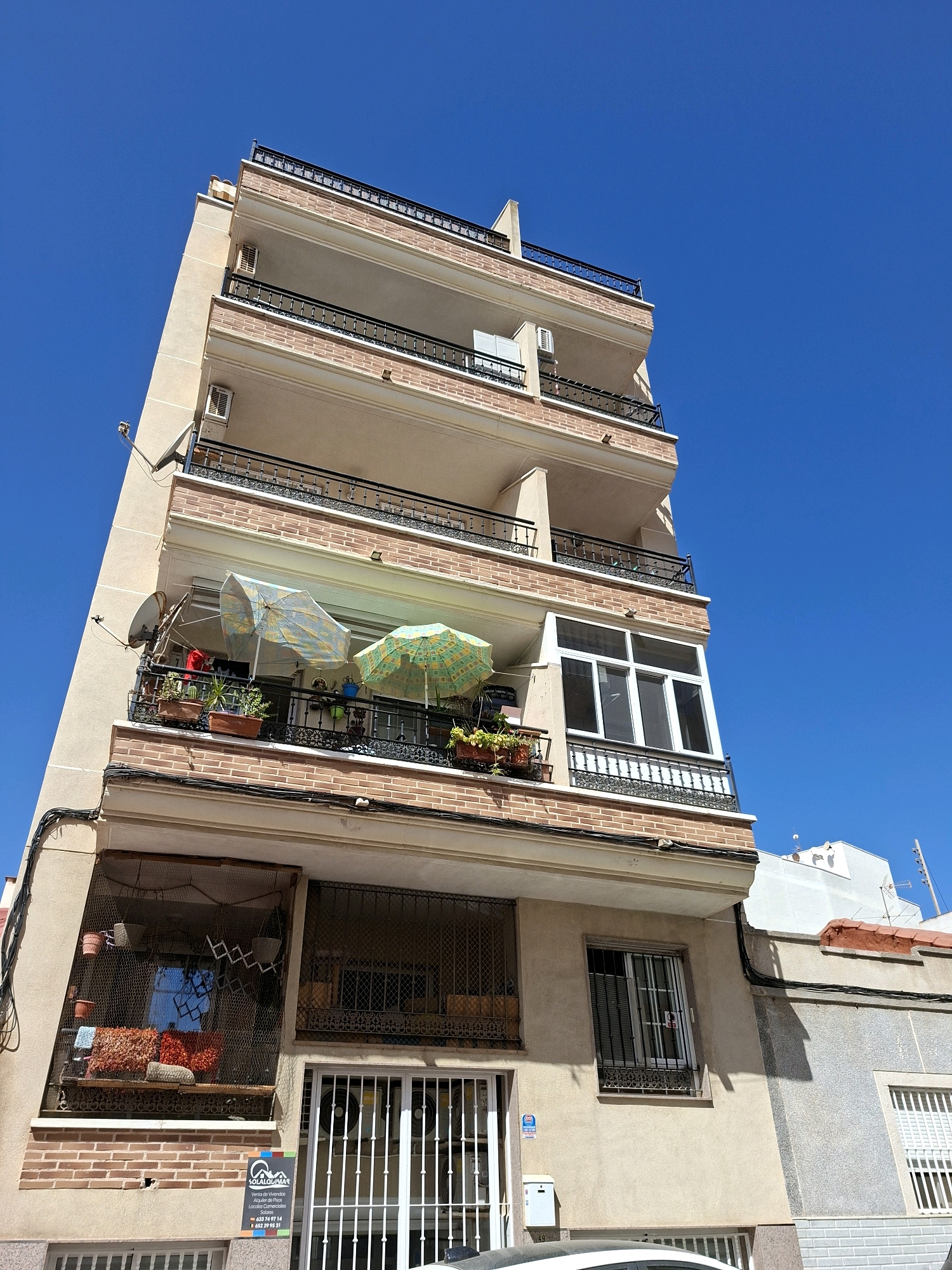 The image shows a multi-story building in Torrevieja, Spain, under a clear blue sky. The building has a pale beige facade with decorative brick accents. There are multiple balconies with black metal railings. Some balconies have potted plants and two are shaded by open umbrellas. The building also features several windows, some with metal grates. The ground floor has a barred window and a white metal gate. A real estate sign is visible on the lower part of the building. The overall scene is typical of a residential building in a Mediterranean coastal city.