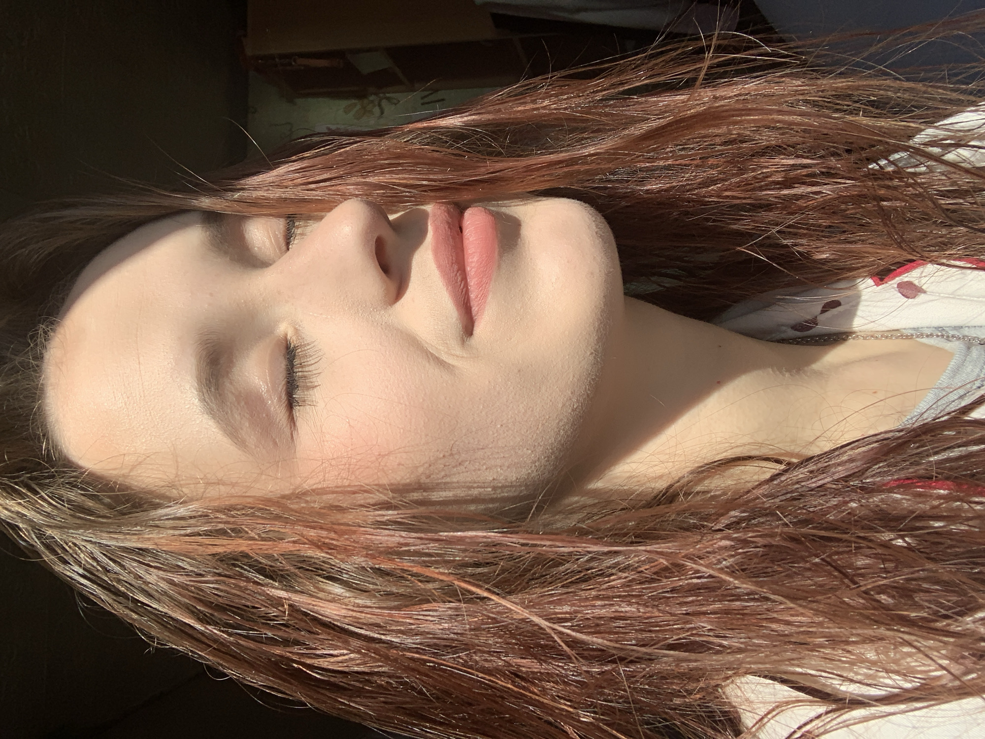 The image captures a close-up, eye-level perspective of a person, likely a young woman, resting. Her head is tilted upwards and to the right, with her eyes gently closed, suggesting sleep or profound relaxation. Strong, warm sunlight illuminates her face and hair, creating distinct highlights and shadows.
The subject has fair, smooth skin and appears to have a light blush on her cheeks. Her lips are full and adorned with a soft pink or nude lipstick. Long, reddish-brown hair, appearing somewhat wet or glossy, is fanned out around her head and shoulders, catching the light in various strands. Visible eyelashes frame her closed eyes, and her eyebrows are naturally shaped.
She is wearing a light-colored top or nightwear with a faint, possibly floral or abstract, red pattern visible on the fabric near her right shoulder. A delicate silver chain necklace is also visible resting on her collarbone area. The background is mostly dark and out of focus, suggesting an indoor setting, possibly a room, but specific details are indistinct.
The strong, directional sunlight indicates either early morning or late afternoon, creating a serene and tranquil atmosphere. The overall impression is one of peaceful rest, bathed in natural light. There is no visible text in the image.