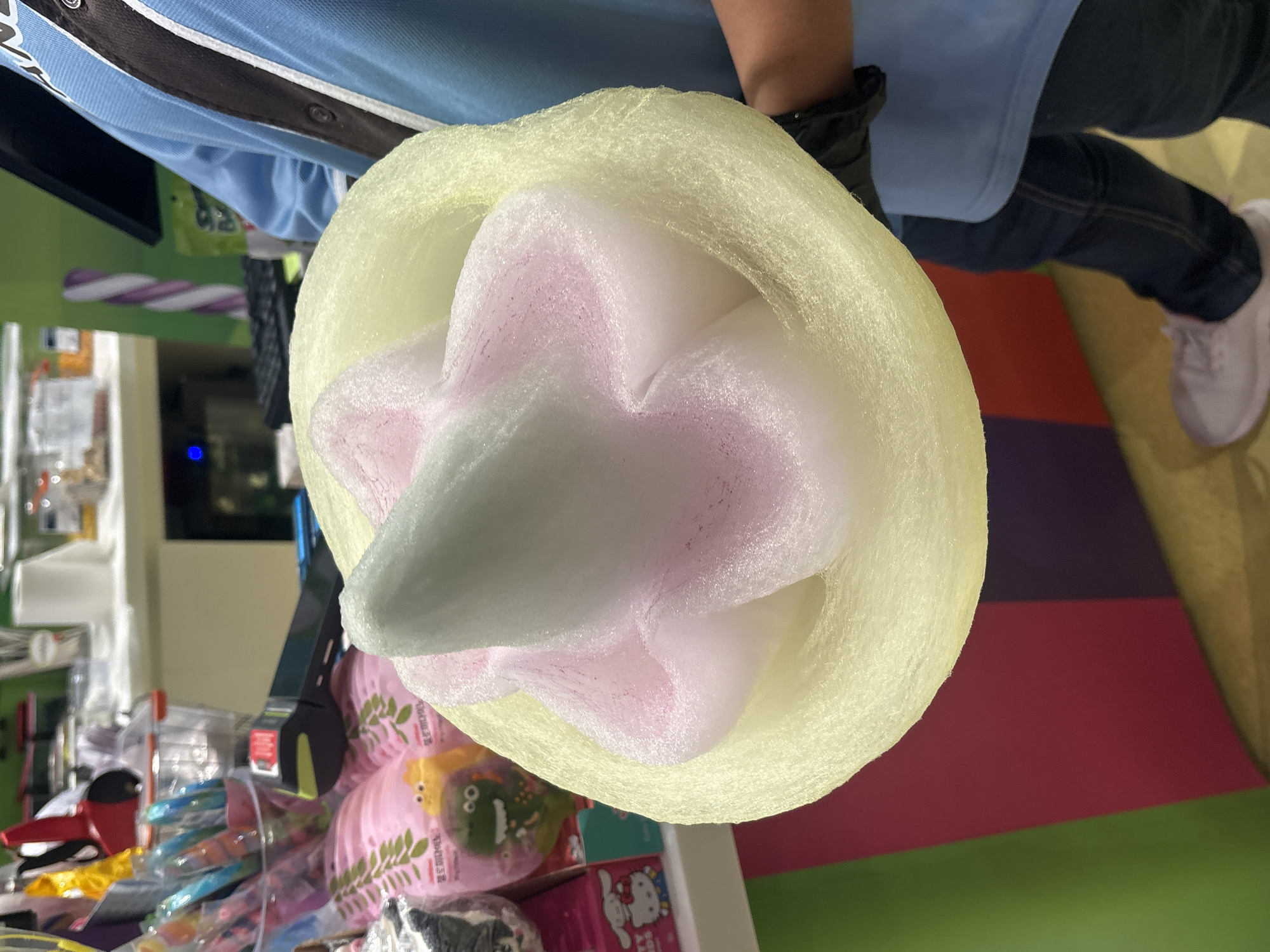 A close-up, overhead shot shows a person holding a large, elaborate piece of pale yellow and pink cotton candy in the shape of a flower. The cotton candy has a light blue, cone-shaped center. The person is wearing a blue shirt with a dark stripe down the front, dark blue jeans, and light pink sneakers. They are also wearing black gloves. In the blurred background, there are shelves stocked with colorful candies and toys. The scene appears to be inside a retail store, possibly a candy shop, with brightly colored vertical panels visible on a wall. This image was taken in Dubai, United Arab Emirates, likely within a commercial establishment catering to sweets and novelty items.