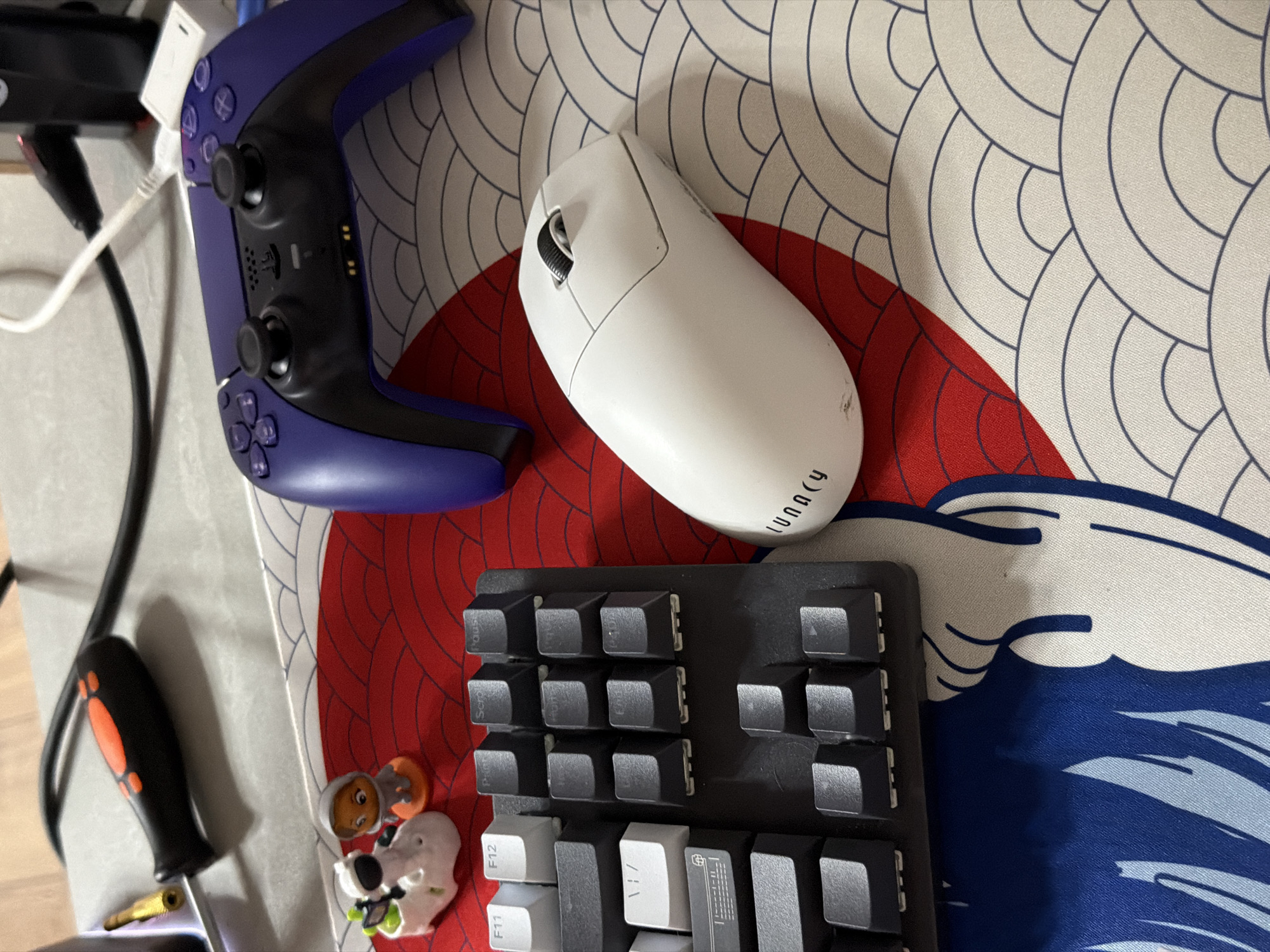A close-up overhead shot shows a desk setup with a purple gaming controller, a white wireless mouse, and a black mechanical keyboard. The objects are placed on a desk mat with a Japanese wave pattern in red, white, and blue. A small screwdriver with an orange and black handle is visible in the upper left corner. There are also two small figurines, one appearing to be a white sheep with green accents and the other a small orange figure in a gray hooded outfit, situated near the top left of the keyboard. The background shows a gray surface with black and white cables. The image does not provide any information about the location in Moscow, Russia.
