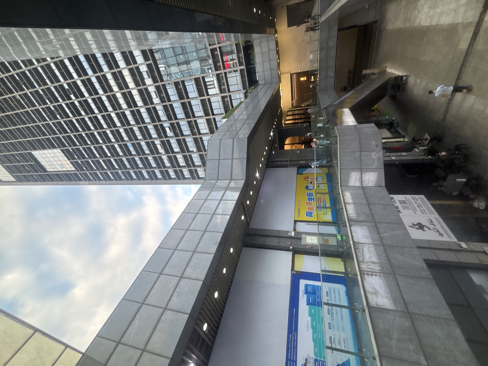 This is an exterior, medium-angle, eye-level shot of a modern commercial building complex in the Liwan District, China. The scene depicts a multi-level structure with exposed concrete and glass elements. In the foreground, a walkway with a glass railing is visible, leading down towards an escalator. People are present on various levels, some walking, others standing. The architecture is characterized by geometric lines and a mix of dark grey concrete and reflective glass surfaces. Sunlight illuminates parts of the scene, creating contrasts and reflections. In the background, a tall, grid-like skyscraper dominates the skyline, with numerous windows. The sky above is partly cloudy. Signs with Chinese characters are visible on the building's facade and on posters.