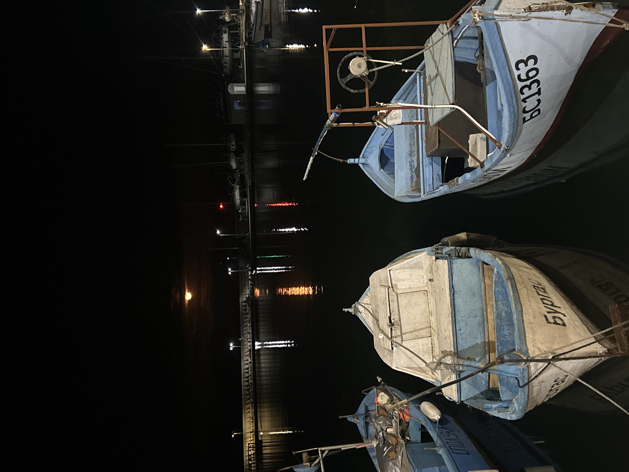 The image shows a nighttime scene at a harbor in Burgas, Bulgaria. Several small fishing boats are docked, with one prominent boat displaying the registration number "БС1363" and another inscribed with the Bulgarian word "Бургас" (Burgas). The harbor is illuminated by streetlights reflected in the water. A pier stretches into the dark water, lined with lights. The overall environment suggests a calm, still night at the harbor.