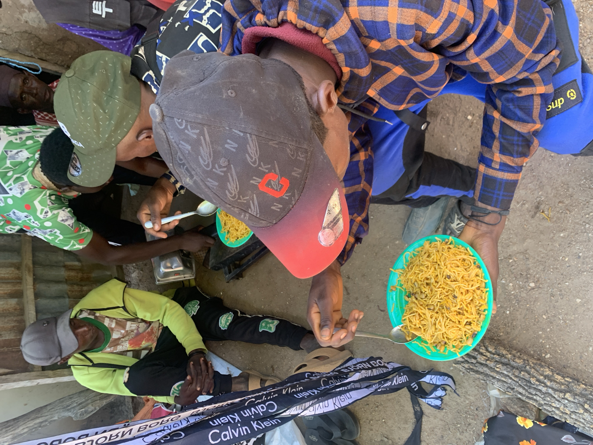 This media file shows a group of people eating together outdoors. The subjects are several individuals, some wearing caps and casual clothing, gathered around to share a meal. One person is holding a plate of noodles and beans with a spoon, in the process of eating. Another person in a green jacket and cap is seated, with their hands clasped. The scene appears to be a casual outdoor gathering, possibly in a rural or less developed area, given the ground surface which is a mix of dirt and gravel, and the presence of what looks like a wooden log.
The activity is clearly eating, suggesting a communal meal or a break for food. The clothing of the individuals indicates a warm climate, likely during the day. One person is wearing a blue and orange plaid shirt, another a green long-sleeved shirt and a grey cap, and another a green shirt with a white pattern.
Visible text includes "RTCAP" on a green cap, and several instances of "Calvin Klein" on a black and white ribbon or fabric strip lying on the ground. There are also some indecipherable characters in what appear to be Asian script on a black bag in the background. The word "Sup" is visible on a yellow and black tag on a blue garment.
The overall impression is one of a simple, communal meal shared in an informal setting. There are no strong cues about emotions, but the act of sharing food often implies togetherness and a sense of community. The location context provided places the scene in Boi A, Nigeria.