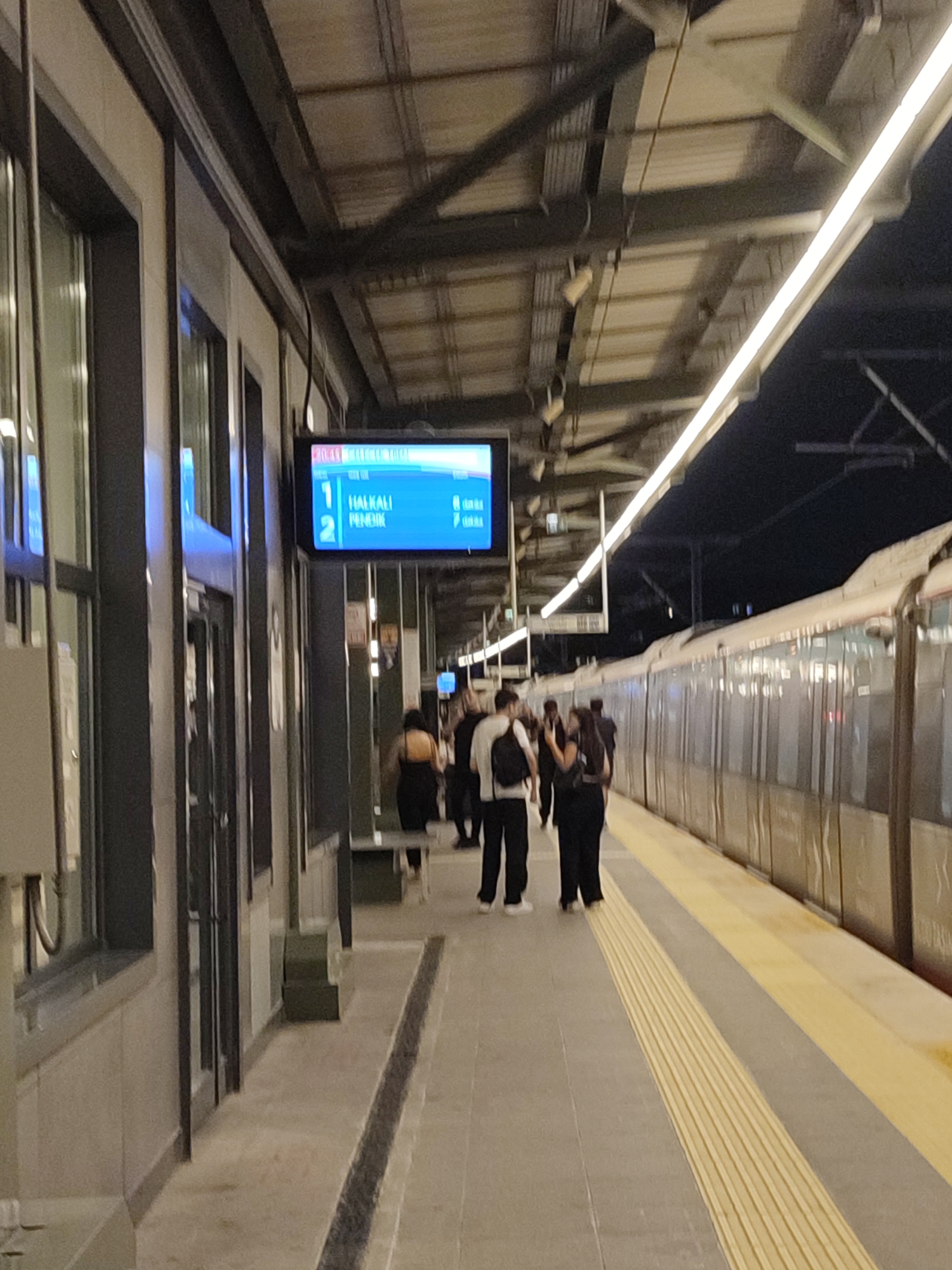 The image depicts a train station platform in Kadıköy, Turkey, at night. A train is stopped at the platform on the right side of the frame. Several individuals are standing on the platform, some facing the train, others in conversation. A digital display board is visible on the left side of the image, showing train destinations and times. The platform is illuminated by overhead lights. The architecture of the station is modern, with exposed beams and metallic structures.