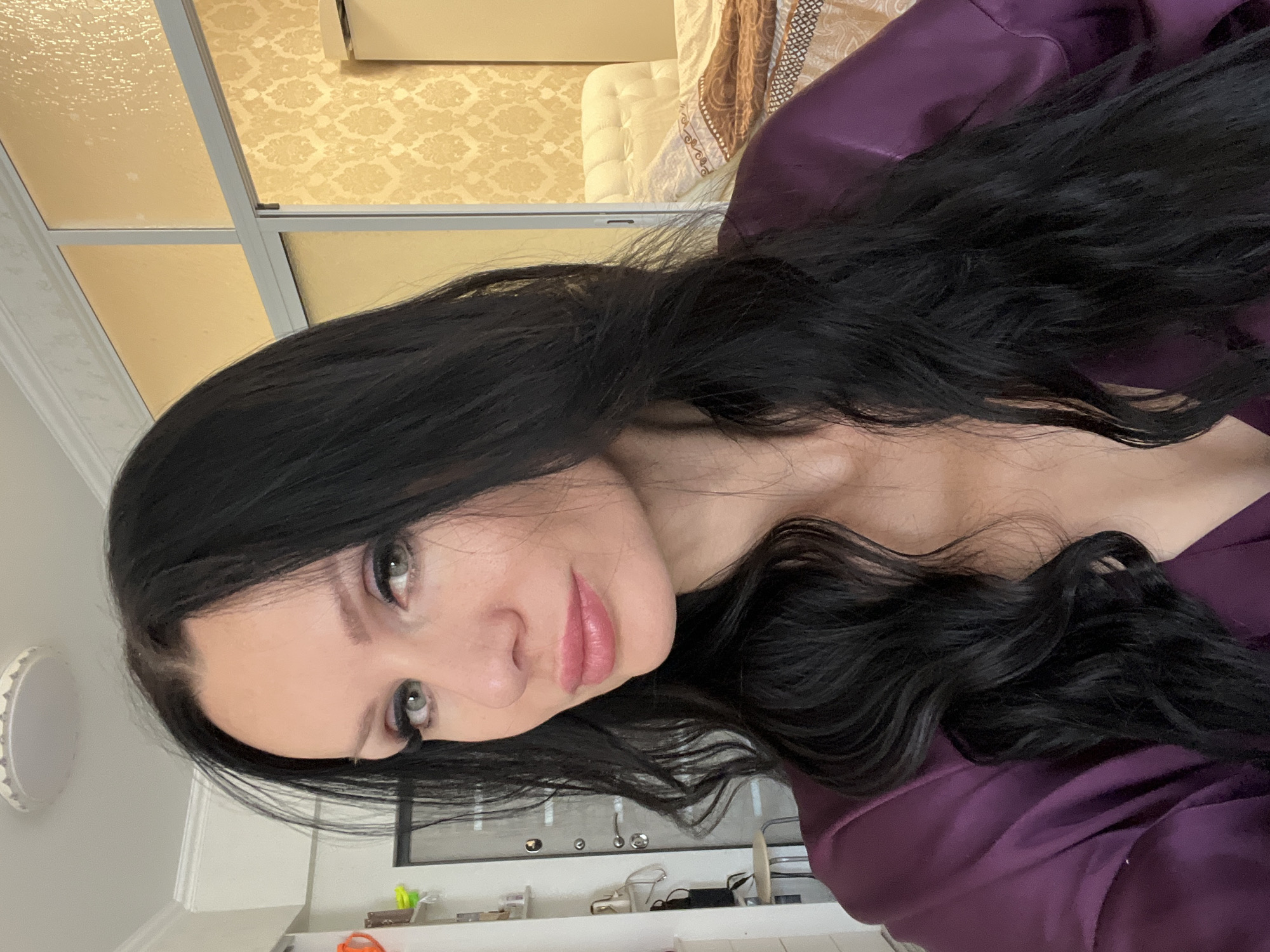 The image shows a medium shot of a woman with long, dark, wavy hair and light-colored eyes. She is wearing a dark purple satin robe. She is looking towards the camera with a slightly parted mouth and subtle makeup. The background is slightly out of focus but appears to be an indoor setting, possibly a bedroom, with light-colored walls, a door, and what looks like a bed with a headboard. There is no indication of the city of Sochi, Russia.