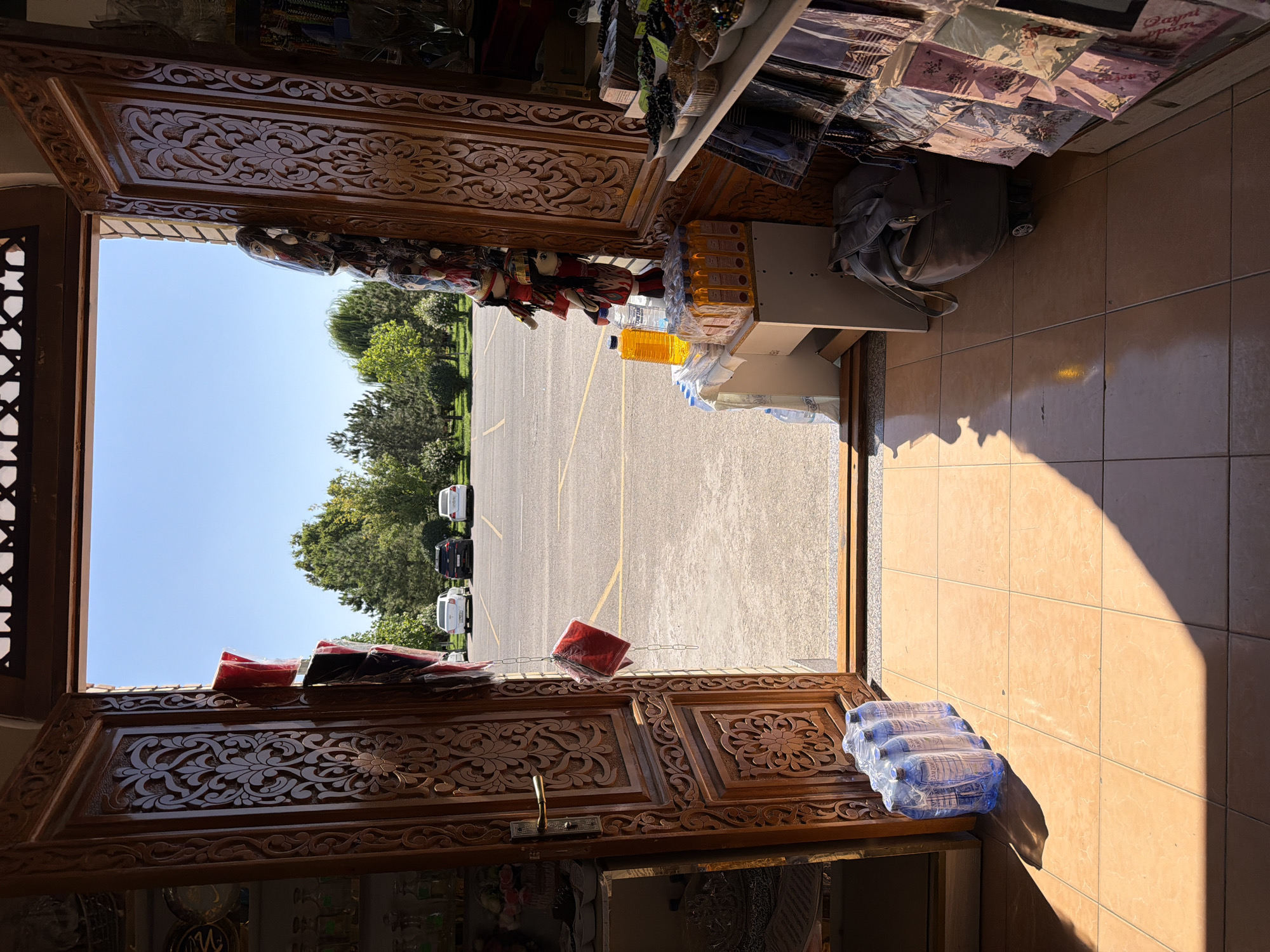 An open doorway with intricately carved wooden doors reveals a bright, sunny outdoor scene. The doorway looks out onto a paved parking lot with several cars parked, including white sedans and a black SUV. Beyond the parking lot, a line of green trees forms a backdrop against a clear blue sky. On the right side of the doorway, inside the building, shelves are stocked with various items, including what appear to be beads, scarves, and other small goods. On the left side, a shelf displays bottles and other products. Several bottles of water are placed on the floor in front of the left door, and a stack of orange liquid bottles sits on a small white table inside the doorway. A suitcase with wheels is also visible on the floor to the right of the small table. The floor inside the doorway is tiled in a warm, sandy color. The overall impression is of a shop opening onto a sunlit parking area in Zangiata, Uzbekistan.