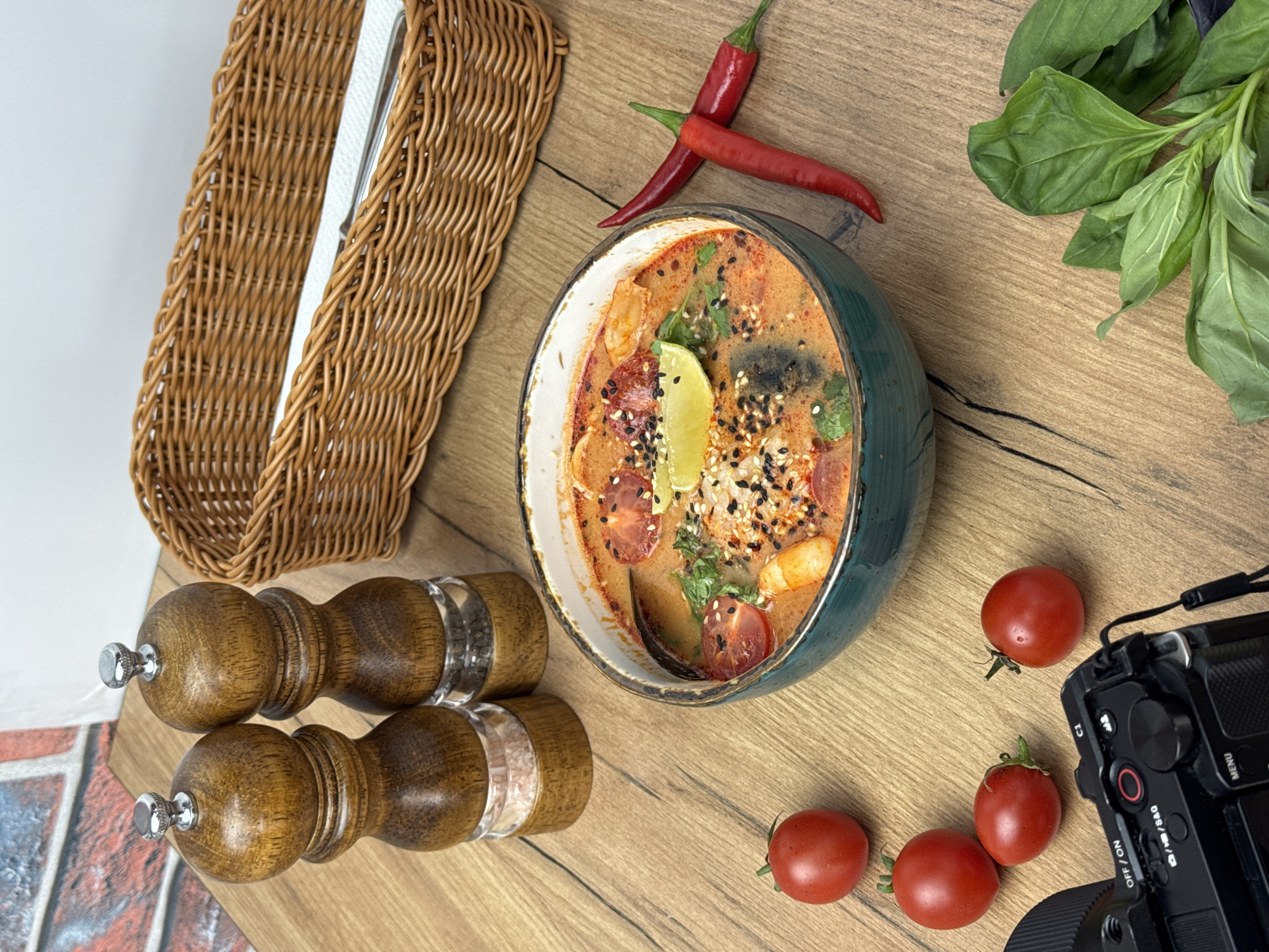 A top-down shot presents a meticulously arranged culinary scene on a light-colored wooden surface. Central to the composition is a green ceramic bowl filled with a rich, red-tinged soup garnished with lime, tomatoes, and herbs, sprinkled with black sesame seeds. Two bright red chili peppers are placed near the bowl. Fresh green basil leaves are arranged on the top right of the frame.
To the left, a brown wicker basket containing white napkins rests. Below the basket are two wooden salt and pepper grinders. Scattered near the lower-right corner of the bowl are four bright red cherry tomatoes. A portion of a black digital camera is visible at the edge.
The scene hints at a setting in Sochi, Russia, owing to the culinary presentation. The photograph appears to be taken indoors with diffused natural light.