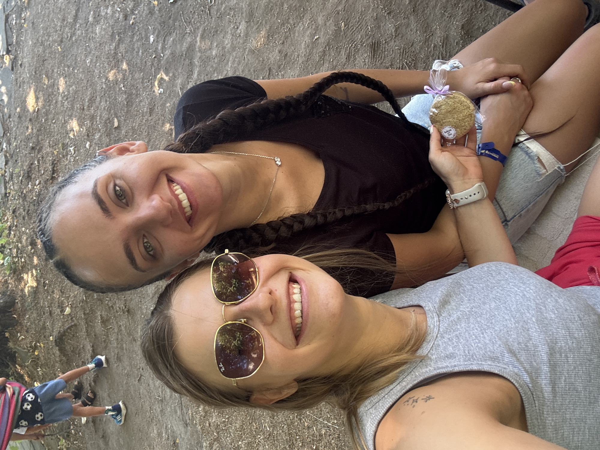 Two young women are sitting outdoors on what appears to be a dusty, unpaved area. The woman on the left is wearing a grey tank top and red shorts, with gold-rimmed sunglasses. She has blonde hair and a tattoo on her left shoulder. She is smiling and holding a cookie wrapped in plastic and tied with a purple ribbon. She also wears a white smartwatch and a blue wristband. The woman on the right is wearing a black t-shirt and denim shorts. She has dark hair styled in two braids. She is also smiling and has a silver necklace with two pendants. In the background, a child is visible wearing blue shorts with soccer ball patterns and a pink and blue sling. The environment is informal, suggesting a casual gathering or event. The overall atmosphere is cheerful. The location is identified as Zheravna, Bulgaria.