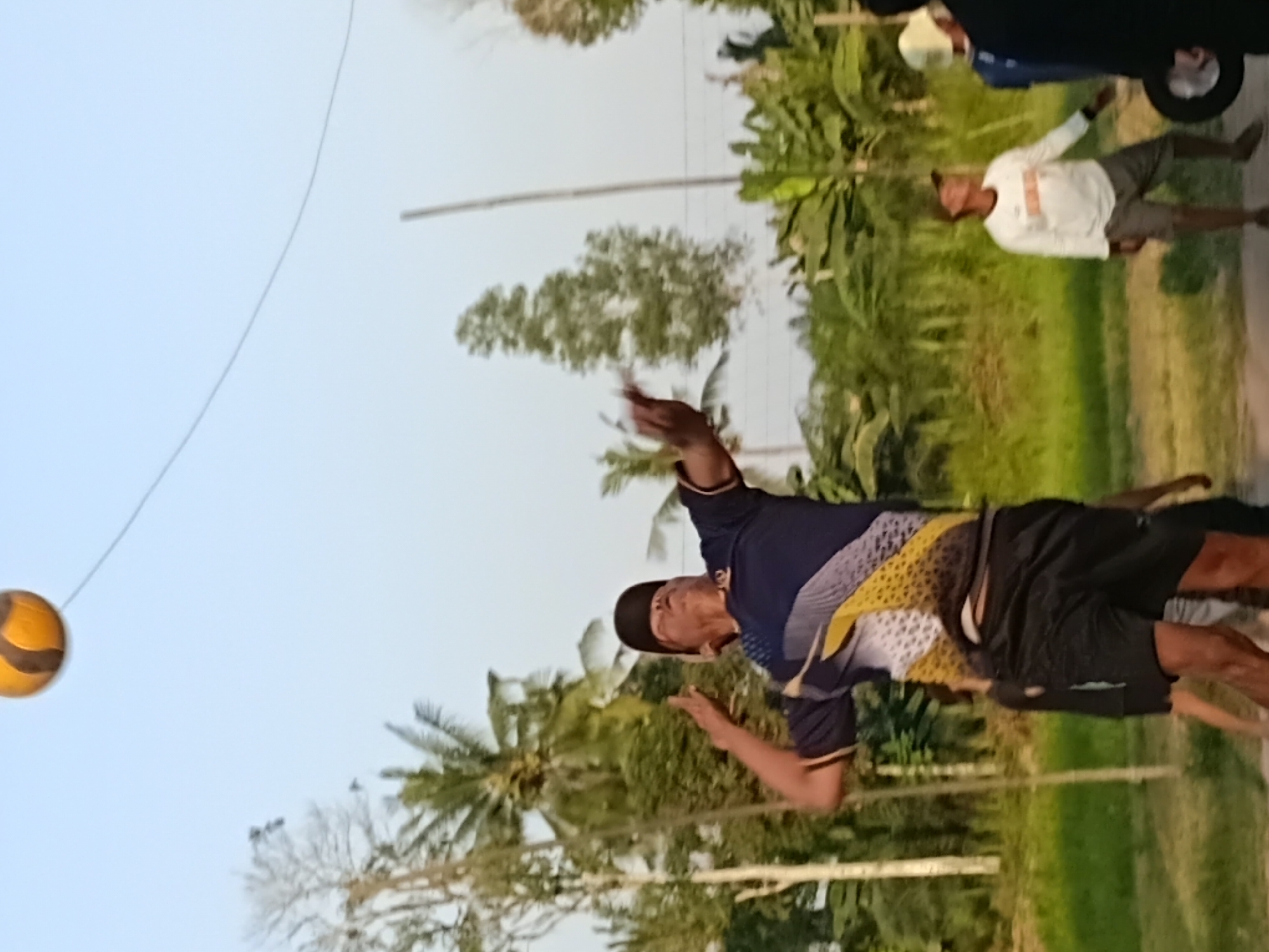 In Margo Rukun, Indonesia, an outdoor volleyball game is in progress. The central focus is a man mid-jump, about to hit a volleyball. He is wearing a dark blue t-shirt with a yellow and white patterned design on the front and black shorts. A dark baseball cap is on his head. In the background, other individuals are visible, including one person in a white long-sleeved shirt and another in a blue shirt. The environment suggests a rural setting with numerous palm trees and dense foliage indicating a tropical climate. The sky is clear and pale blue.