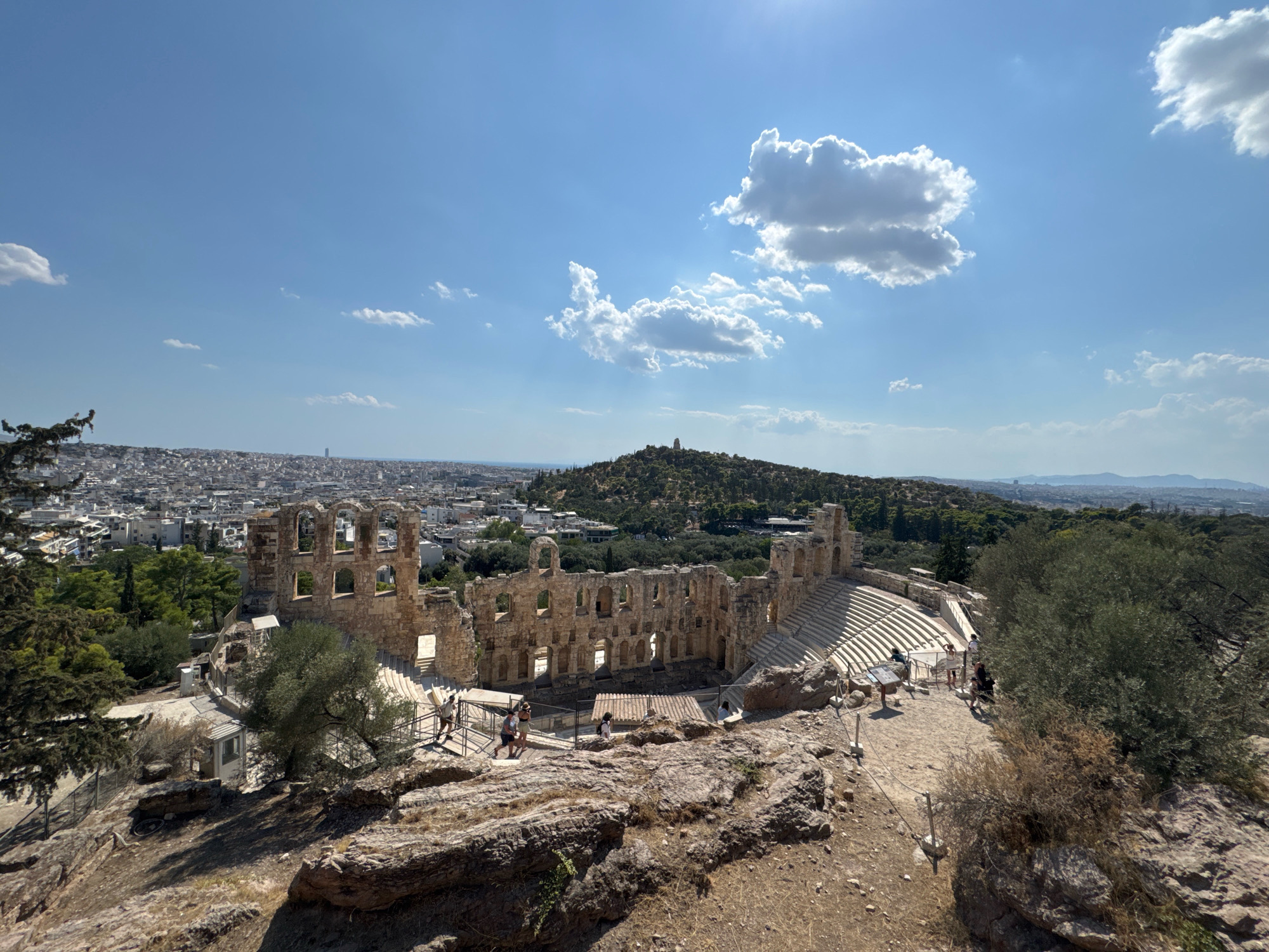 The image captures an outdoor panoramic view of the Odeon of Herodes Atticus, a historic stone amphitheater located on the southwest slope of the Acropolis of Athens, Greece. The ancient structure, characterized by its tiered seating and weathered stone walls with arched openings, occupies the central foreground. In front of the amphitheater, several individuals are visible, some walking along pathways and others standing near railings, appearing to be tourists or visitors. The foreground consists of rocky terrain with scattered dry vegetation and trees. Beyond the amphitheater, the cityscape of Athens stretches out, with numerous buildings and a distant hazy horizon under a bright, sunny sky dotted with scattered white clouds. The overall scene depicts a clear, warm day at a significant historical landmark within an urban setting.