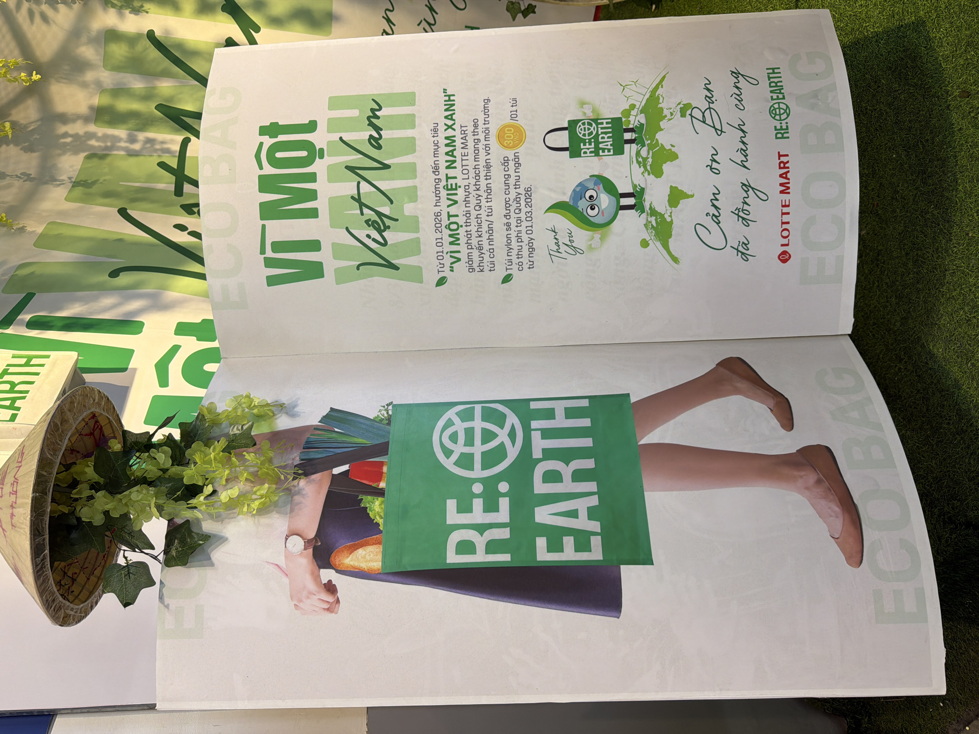 The image is a close-up shot of an open brochure or poster featuring environmental and sustainability messages, presumably from LOTTE MART. The brochure is entitled "Vì Một Việt Nam Xanh," which translates to "For a Green Vietnam."
On the left page, there's an image of a person holding a green shopping bag with the words "RE: EARTH" on it. The bag seems to be filled with food items. A traditional Vietnamese hat is placed next to the brochure, with some greenery.
The right page of the brochure provides details of an initiative, stating that from January 1, 2026, LOTTE MART encourages customers to bring their own reusable bags to reduce plastic waste. Nylon bags will be provided at a cost of 300 VND per bag at the checkout from March 1, 2026. The page also includes graphics with an environmental theme and the message, "Thank You" and "Cảm ơn Bạn đã đồng hành cùng LOTTE MART RE:EARTH" which translates to "Thank you for your partnership with LOTTE MART RE: EARTH".
The text suggests that this advertisement promotes environmental consciousness and sustainability efforts, likely related to reducing plastic usage at LOTTE MART stores in Vietnam. There is no mention of Da Nang specifically.