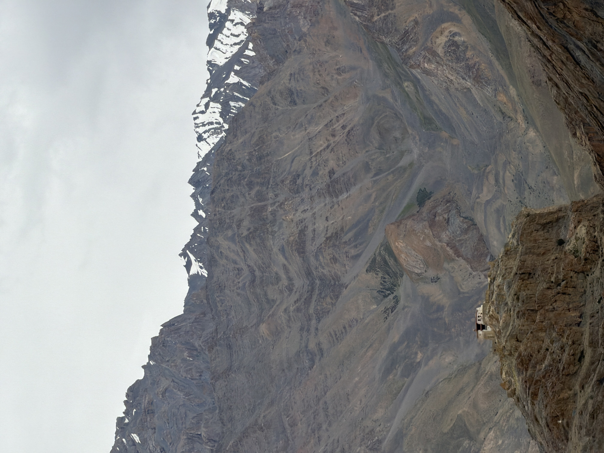 A remote monastery sits atop a rocky outcrop in Zangla, India, under a cloudy sky. The monastery, with its white walls and red roof, is small and appears to be of traditional architecture. It is perched on a steep, rugged hill, surrounded by barren, mountainous terrain. The mountains in the background are imposing, with jagged peaks and slopes covered in scree and rocks. Some of the higher peaks are dusted with snow. The overall impression is one of isolation and grandeur, typical of the high-altitude regions of India.