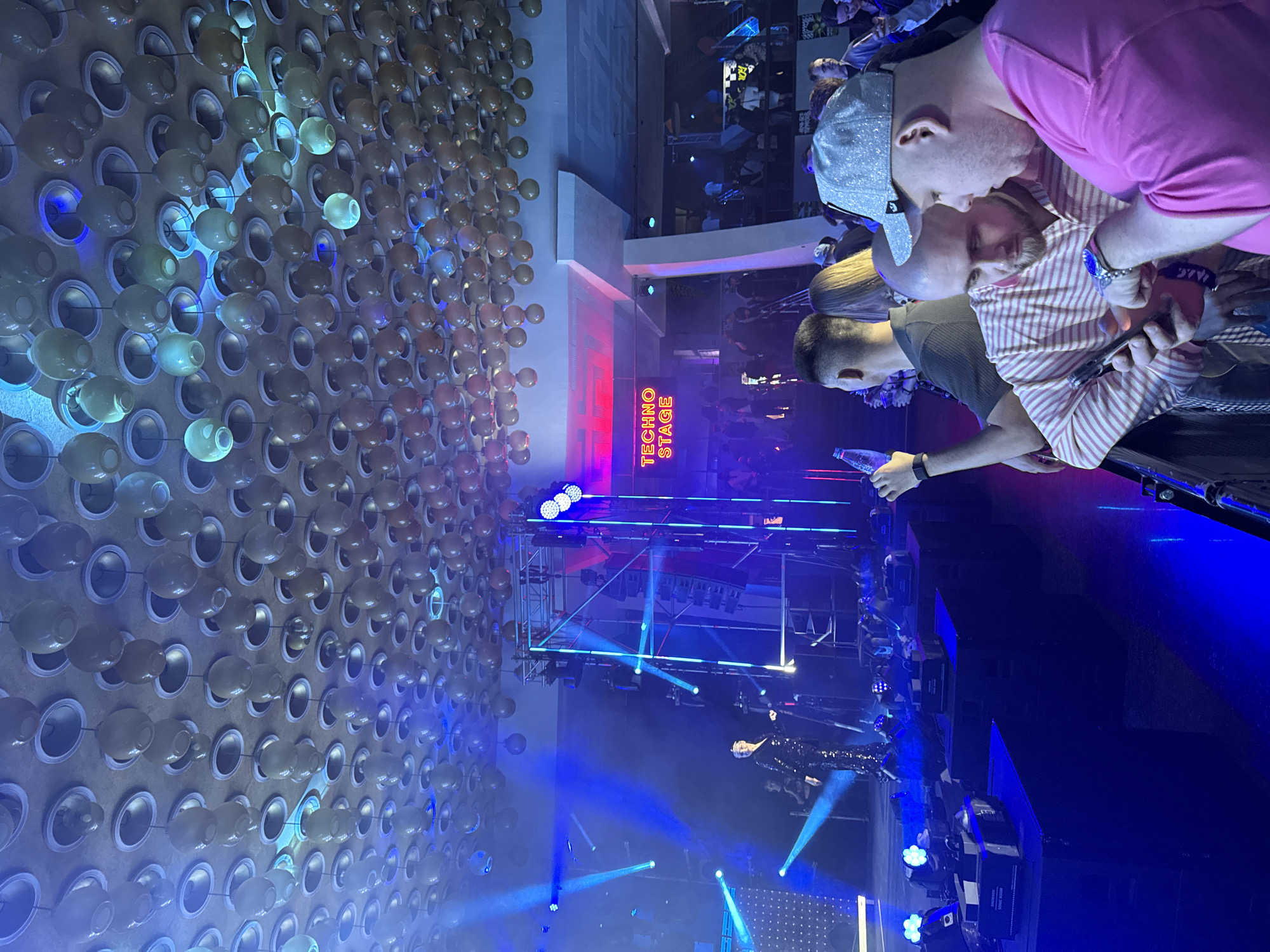 The image depicts a scene at a "Techno Stage" in Yerevan, Armenia. A wall with several rows of small pots or containers is on the left. A stage with lighting equipment and a performer is centered. A group of people are near the stage. Two people are standing close to the photographer in the foreground. One is wearing a pink shirt, and the other is wearing a striped shirt.