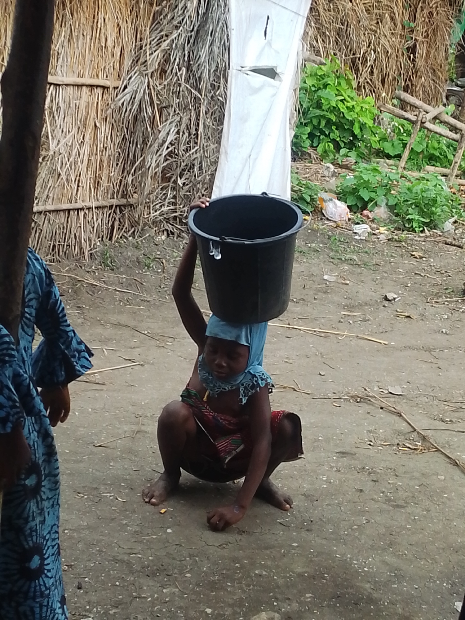 An outdoor scene in Otulu, Nigeria, shows a young child wearing a blue head covering and a colorful patterned garment, squatting and balancing a black plastic bucket on their head. Another person, partially visible on the left, wears a blue patterned dress. The ground is a rough, dusty surface, and the background features a thatched wall, some green foliage, and wooden structures. The overall impression is one of a simple, possibly rural, setting.