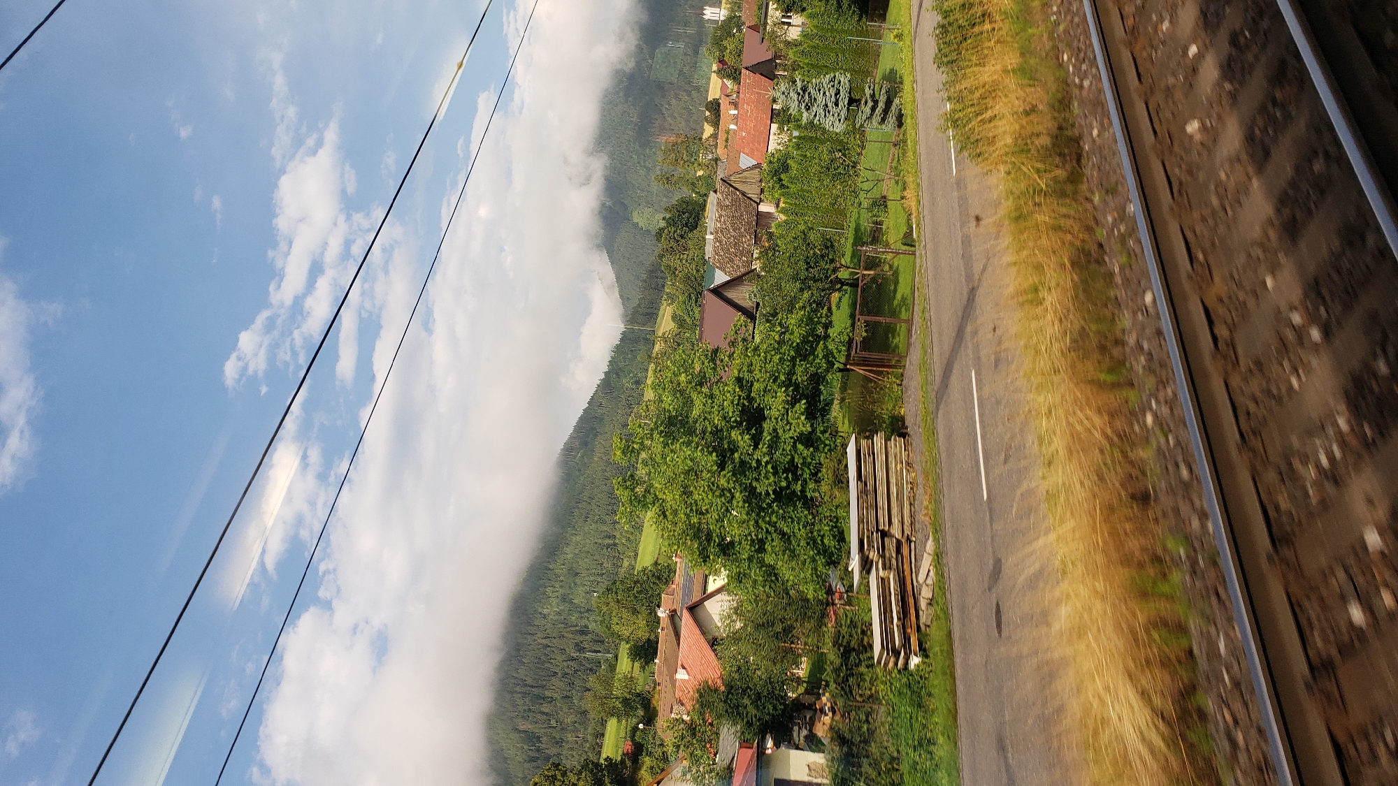 The image depicts a rural village scene in Kráľova Lehota, Slovakia, viewed from a moving train. In the foreground, train tracks run horizontally across the bottom of the frame. Beyond the tracks, a strip of dry, golden grass borders a paved road with a white line marking its center. To the left of the road, a stack of lumber is piled. Further back, a cluster of houses with red-tiled roofs and some with grey shingles are visible amidst lush green trees and manicured lawns. The village is nestled at the foot of a densely forested mountain range, which is partially obscured by low-hanging clouds and mist. The sky above is a clear blue with scattered white clouds, and overhead power lines stretch across the upper portion of the image.