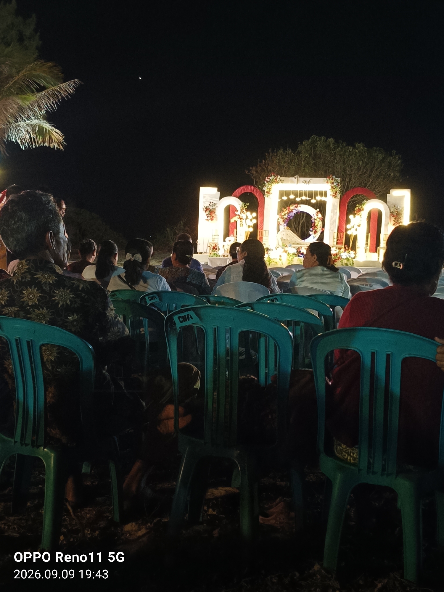 The image depicts an outdoor nighttime event with a decorative archway and illuminated text, likely a wedding or celebration, in Otan, Indonesia. The foreground features several rows of teal plastic chairs occupied by a diverse group of people, their backs mostly to the camera. The individuals are casually dressed, with some sporting traditional attire. In the background, a large, brightly lit archway is adorned with floral arrangements and the letters "M" and "O" are prominently displayed, possibly part of a larger word or monogram. The ground appears to be sand or dirt, suggesting a beach or open-air venue. A palm tree is visible on the far left. The overall atmosphere is festive and communal.