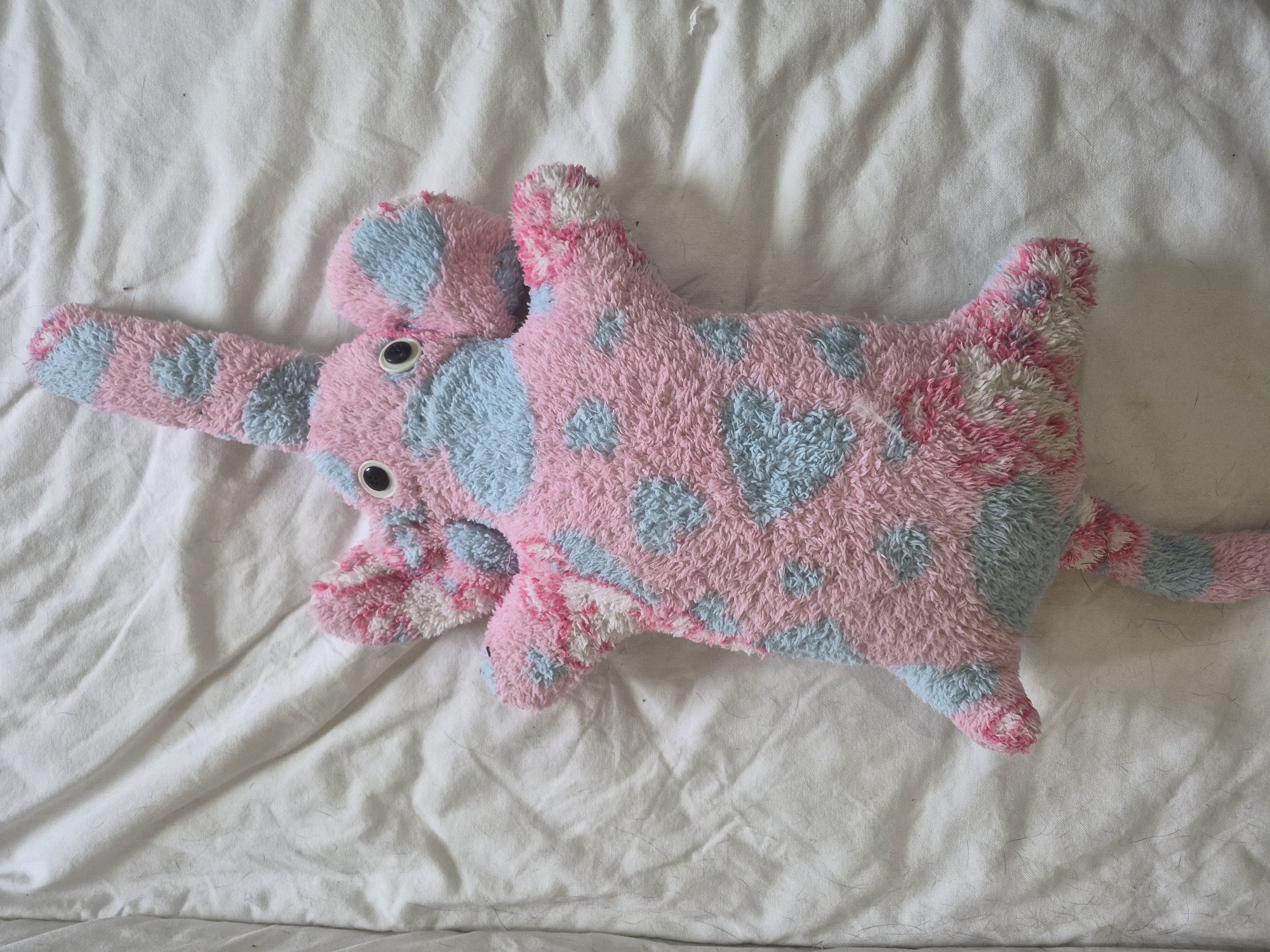 A plush toy elephant, predominantly pink with blue polka dots, lies on a white, wrinkled bedsheet. The elephant has large, googly eyes, floppy ears, and a long trunk. Its body is elongated, and it has a tail. The fabric appears well-worn and slightly matted. The background is a creased white sheet, with visible fibers and dust particles. The location is not specified within Vlast Truda, Russia.