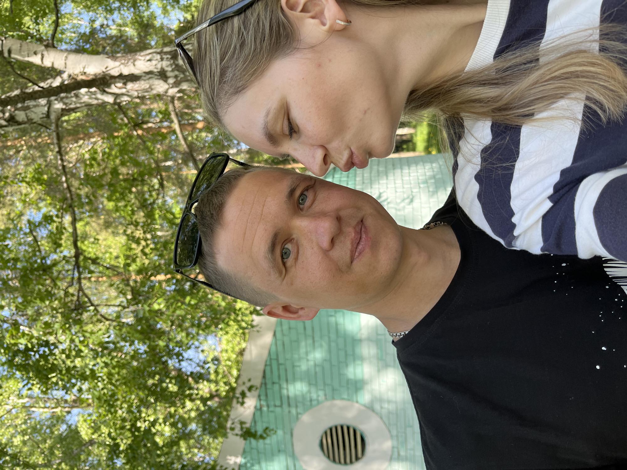 A fair-skinned man and woman pose in a close outdoor shot, with the man wearing a black T-shirt and a silver chain necklace. Sunglasses rest on his head. The woman, with blond hair, wears a white and blue striped shirt and sunglasses perched on her head. Her face is close to the man's, as if she is about to kiss him. The background shows a green brick structure with a white circular vent, partially obscured by tree foliage. The location appears to be a park in the city of Chaikovsky, Russia.