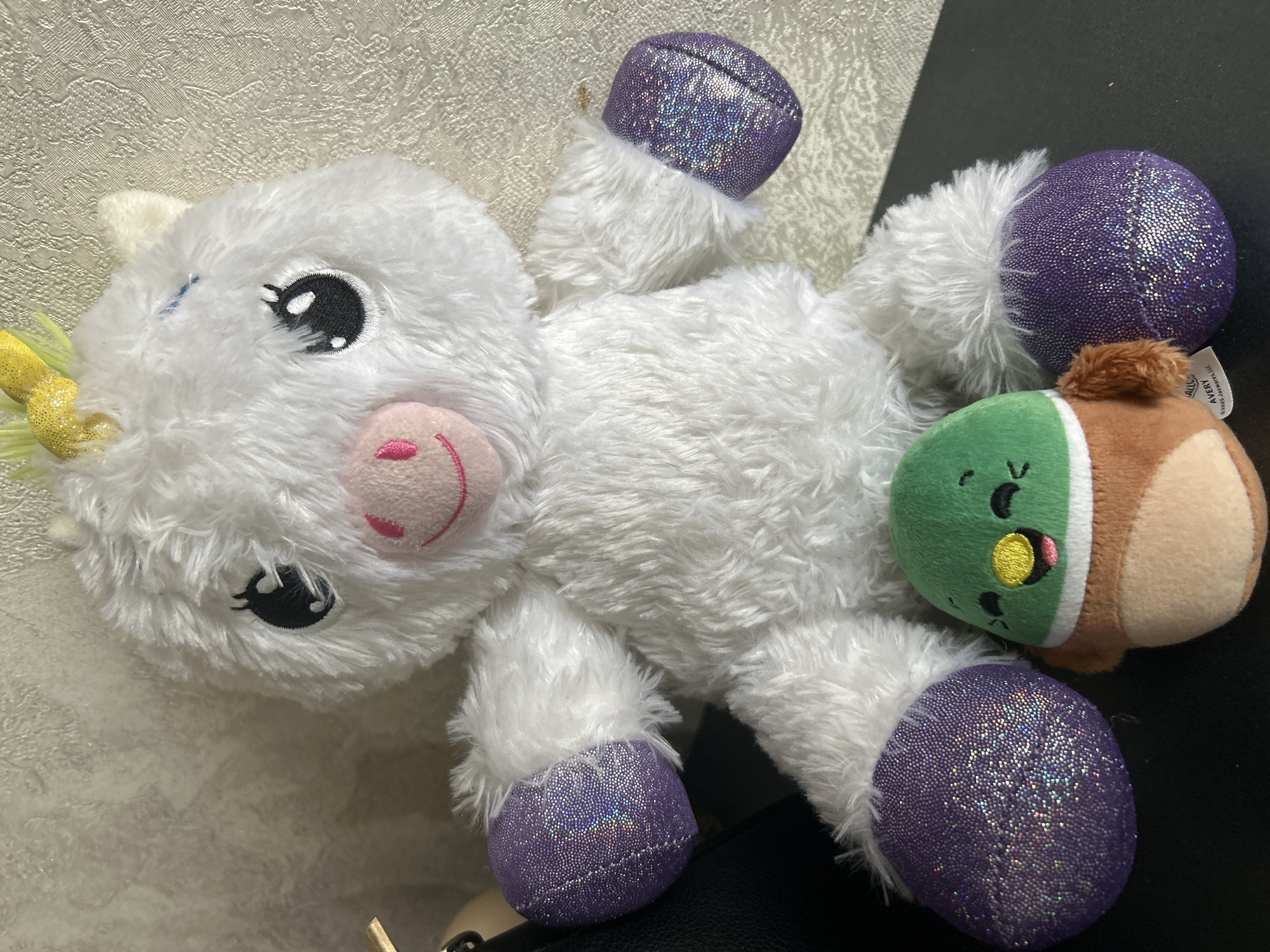 A close-up shot depicts two plush toys: a white unicorn with purple, glittery hooves and a small, round toy resembling a duck or bird in brown and green. The unicorn has a pink smiling mouth and a yellow horn. They are laying on a surface with a light colored textured background, which transitions to a dark surface with a grainy quality. The city of Tbilisi, Georgia, is not discernible from the visual information provided in the image.
