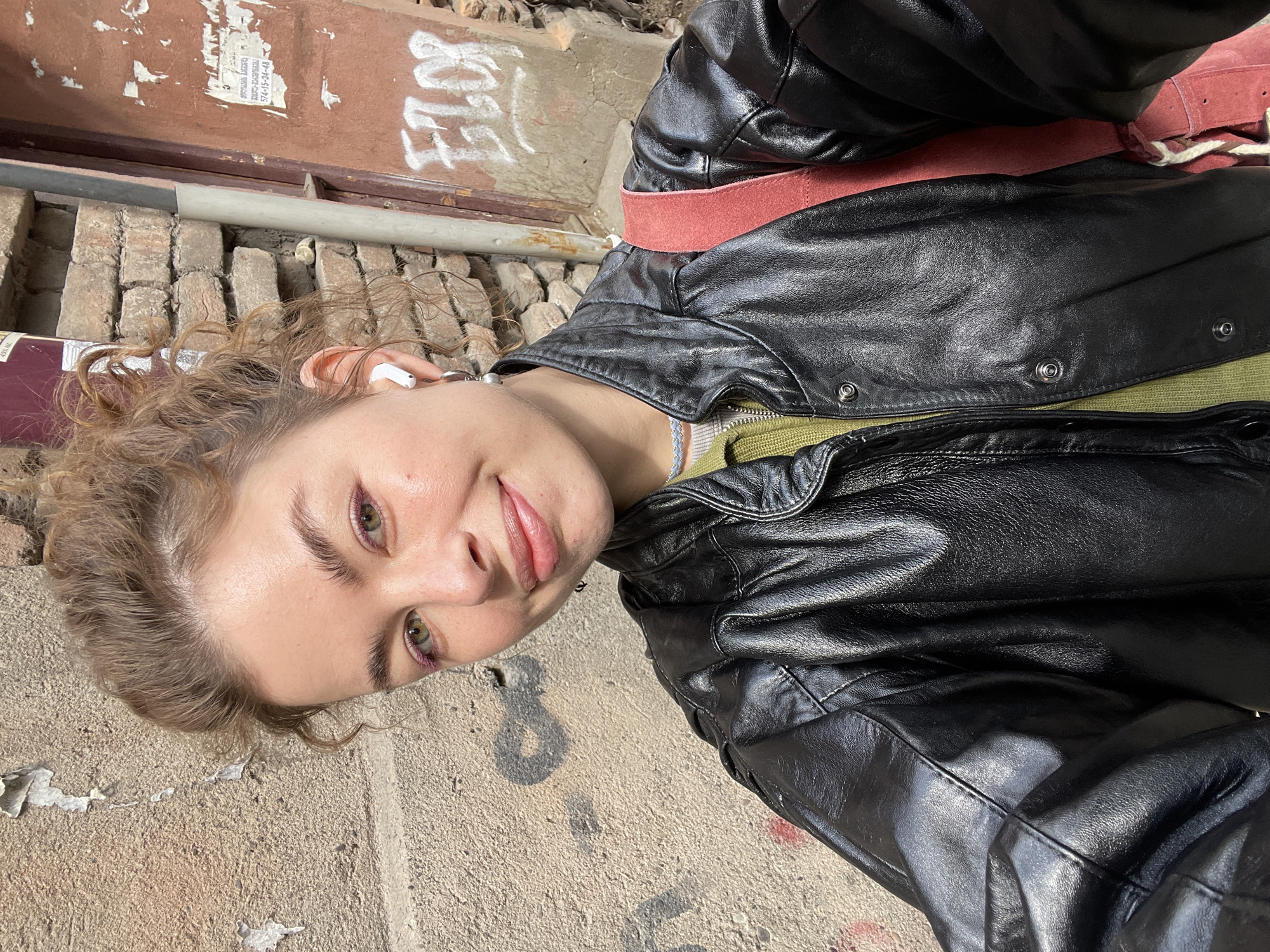 A young, light-skinned woman with curly blonde hair and green eyes is shown in a close-up selfie. She wears a black leather jacket over a green shirt with a glimpse of white underneath. A red belt is visible over the jacket. She has an earbud in her left ear. The background is a city street, possibly in Tbilisi, Georgia, with a concrete sidewalk and a brick wall. Graffiti is visible on the wall and sidewalk.