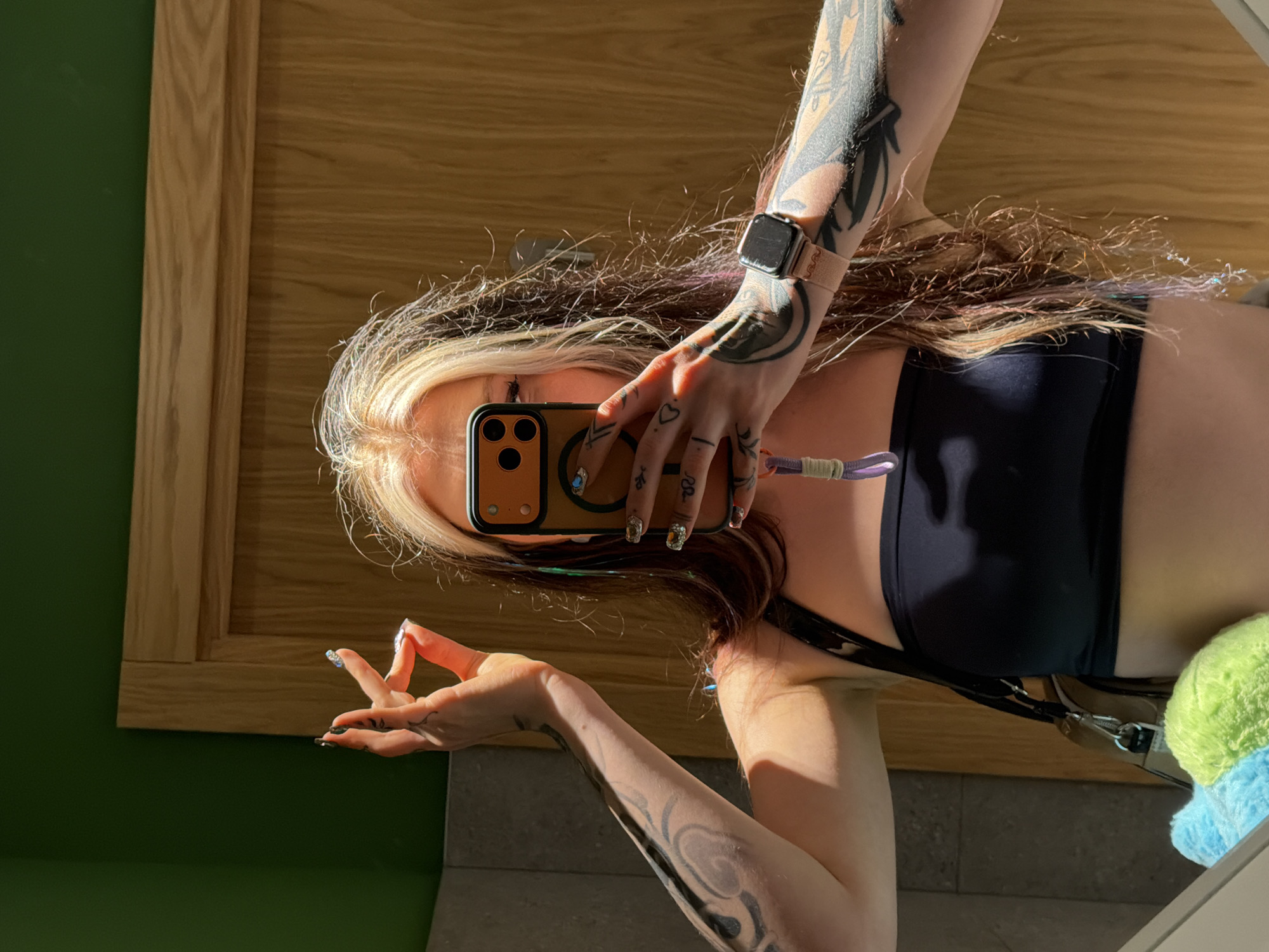A person is taking a selfie in a bathroom. The person is fair-skinned with blonde and brown hair, wearing a black sports bra, and has multiple tattoos on both arms and hands. The person is holding a phone with a brown case, obscuring their face. They are wearing an Apple watch. The background shows a wooden-framed mirror, a dark green wall, and bathroom tiling. The exact location in Belgrade, Serbia, cannot be determined from the image.